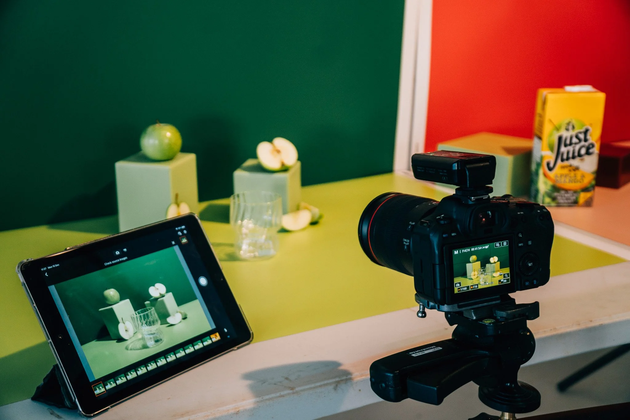 A camera on a tripod capturing a scene of green apples and apple slices on small pedestals, with a juice box in the background, as seen on a tablet and a professional camera screen.