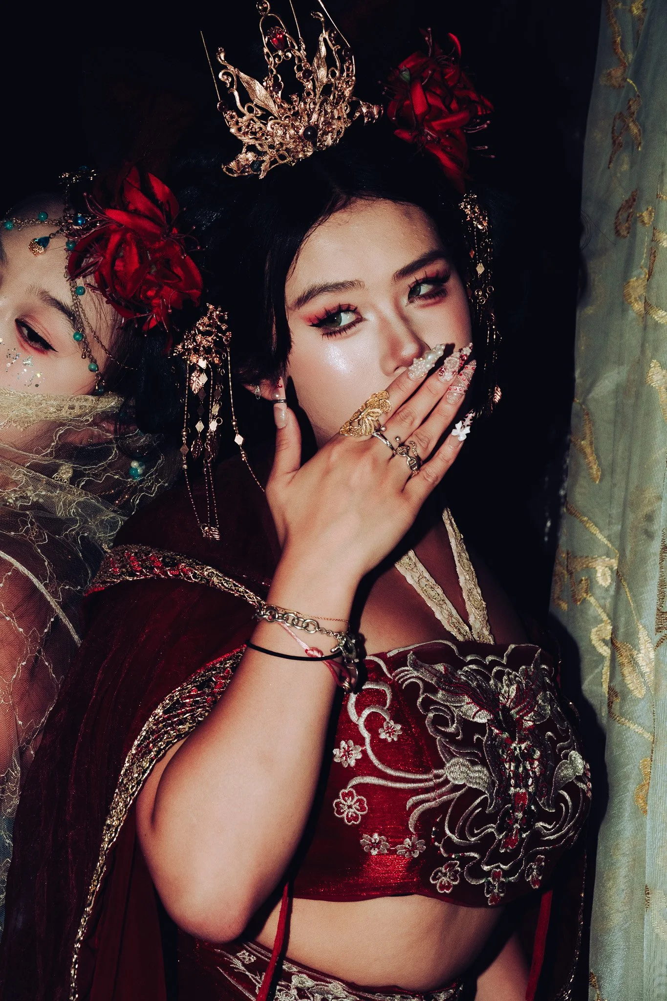 A woman dressed in traditional Asian attire, wearing ornate jewelry and a decorative crown, with dark hair and makeup, partially covering her face with her hand.