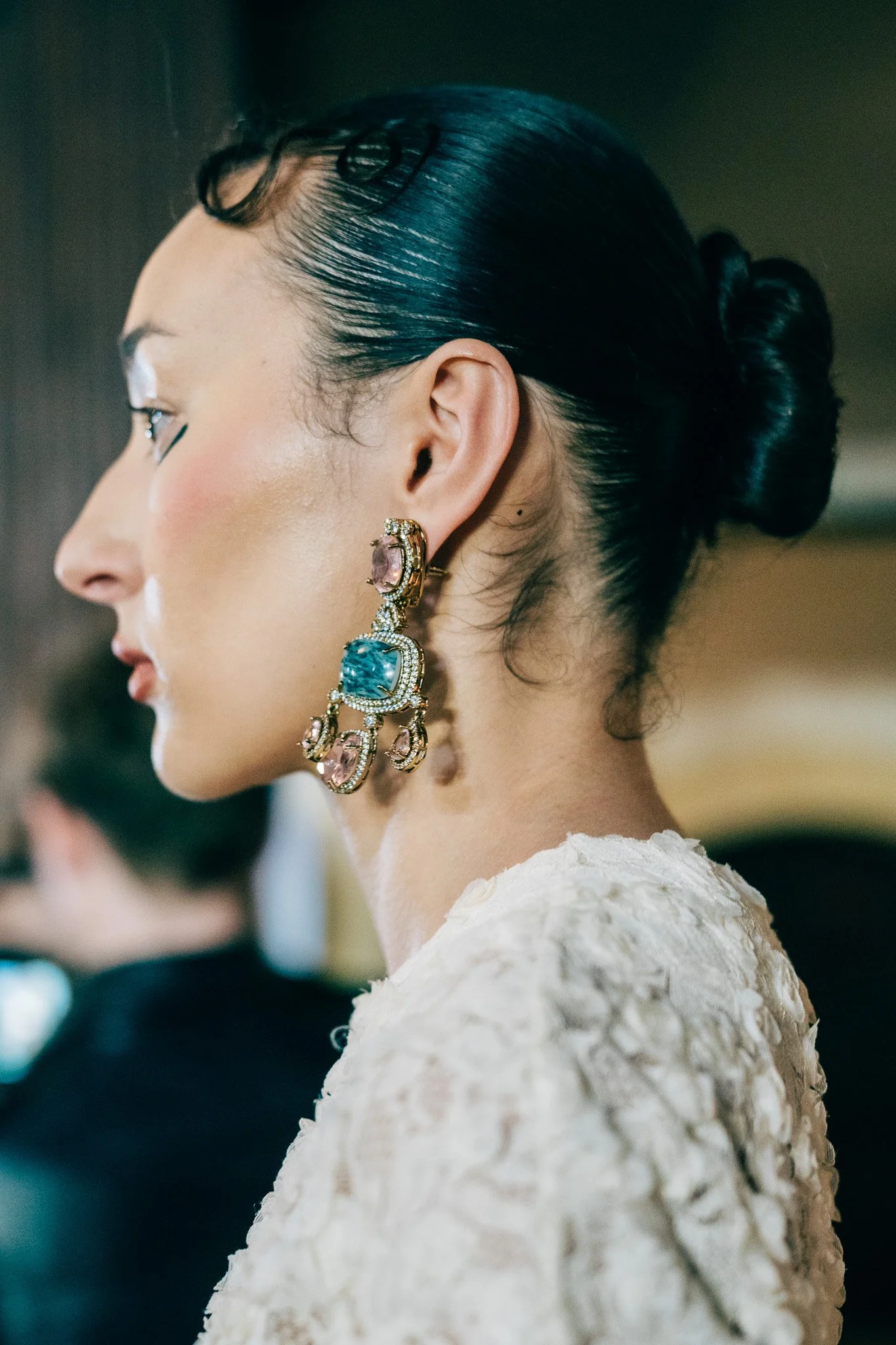 Side profile of a woman with slicked-back black hair styled in an elegant bun, wearing ornate earrings and a white textured dress.