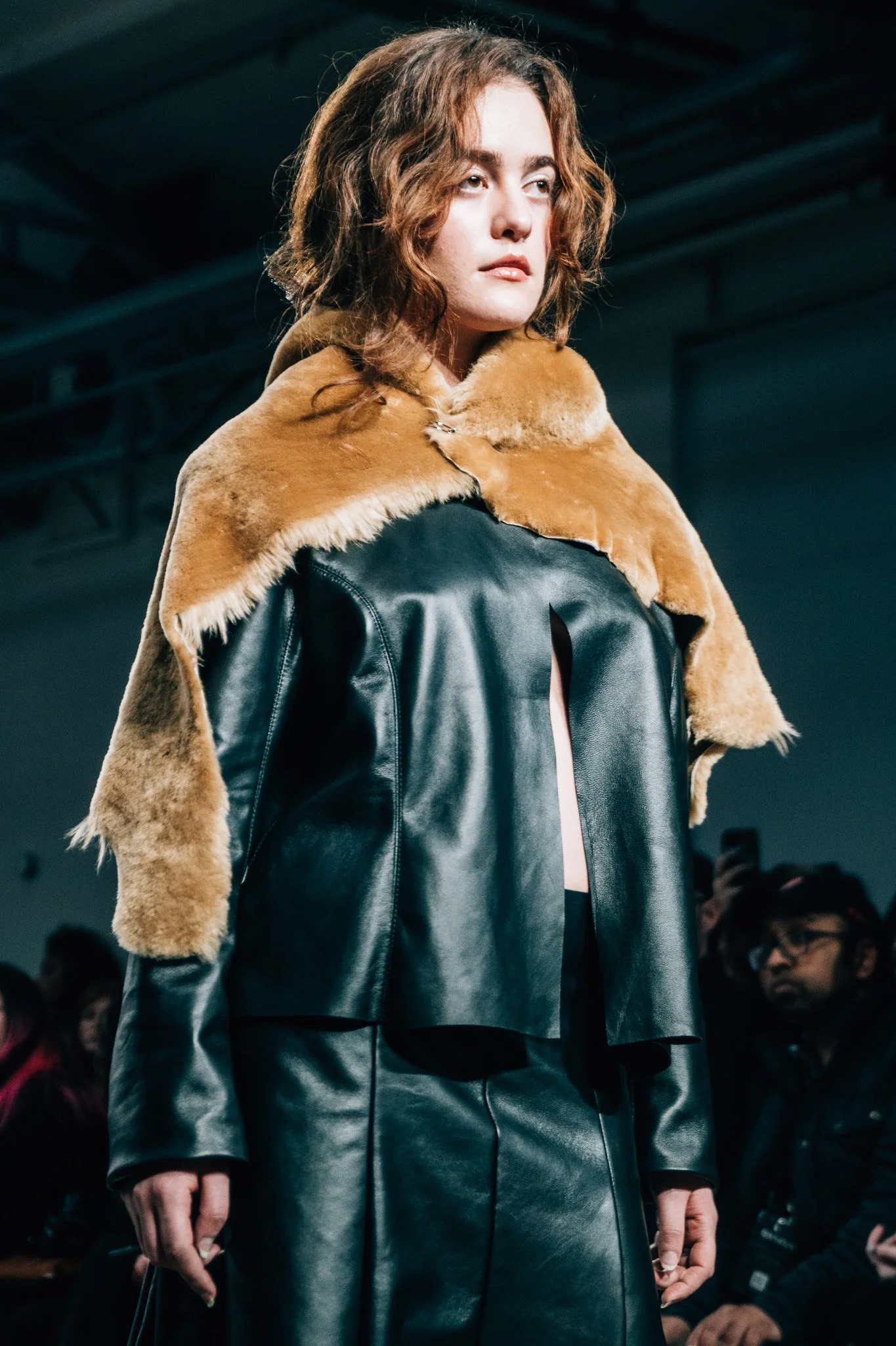 Model walking down runway during fashion show, wearing a black leather coat with tan fur collar.