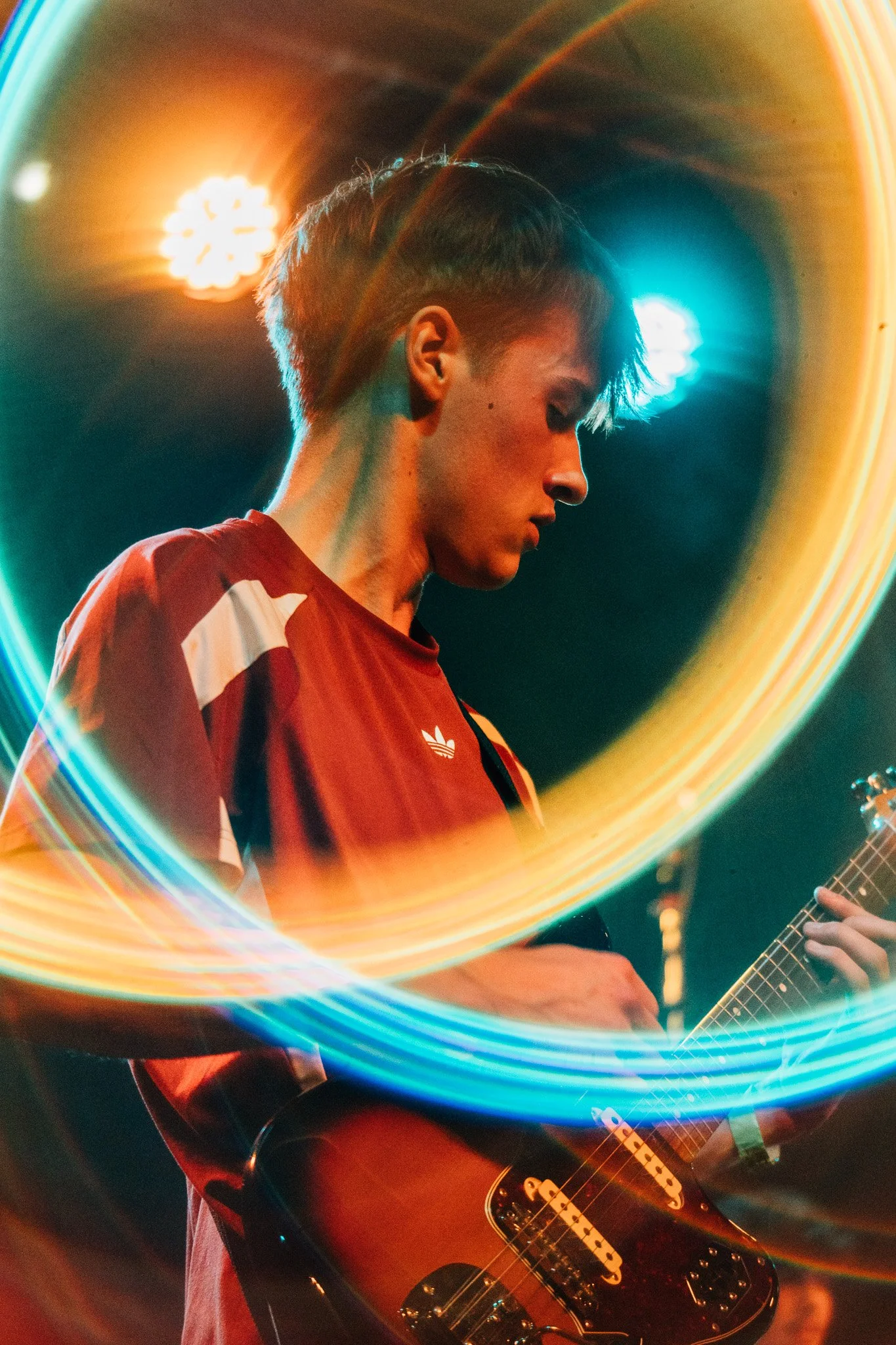 A young man with short hair wearing a red and white shirt playing an electric guitar on stage with colorful lights creating a circular light pattern around him.