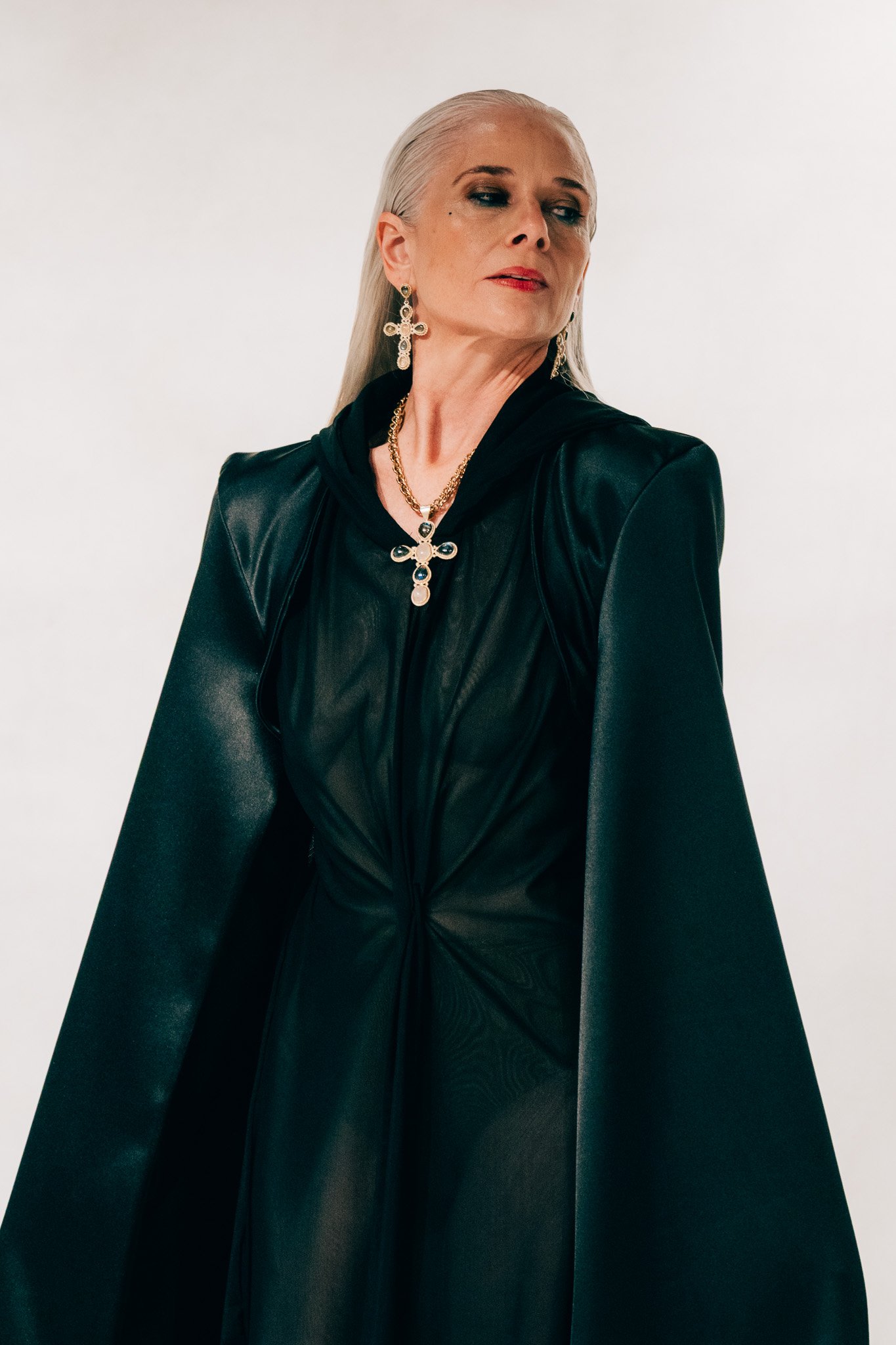 A woman with silver hair, wearing large cross-shaped earrings, a gold chain necklace with a cross pendant, a black leather jacket, and a black top, looking to the side against a neutral background.