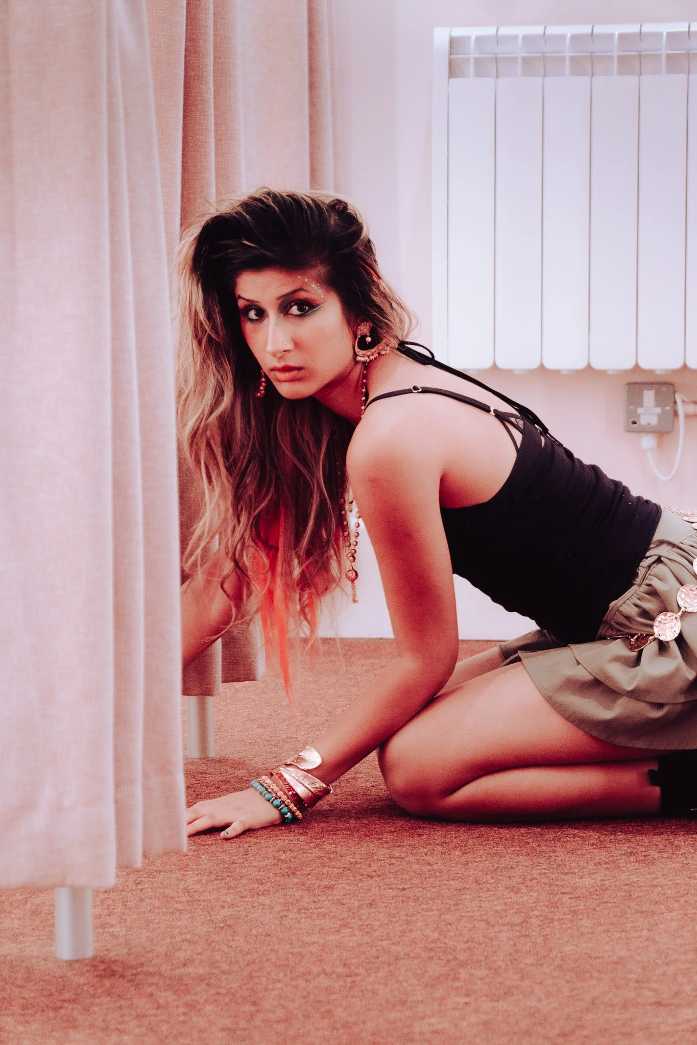 A woman with long wavy hair, wearing earrings, necklaces, bracelets, a black tank top, and khaki shorts, crouches on a carpeted floor near a corner with a radiator and pink curtains.