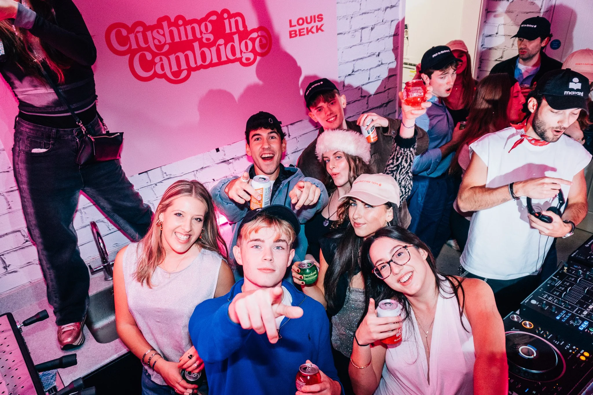 Group of young people at a lively party, smiling, holding drinks, and enjoying music with a DJ in the foreground. A pink wall with a 'Crushing in Cambridge' sign is in the background.
