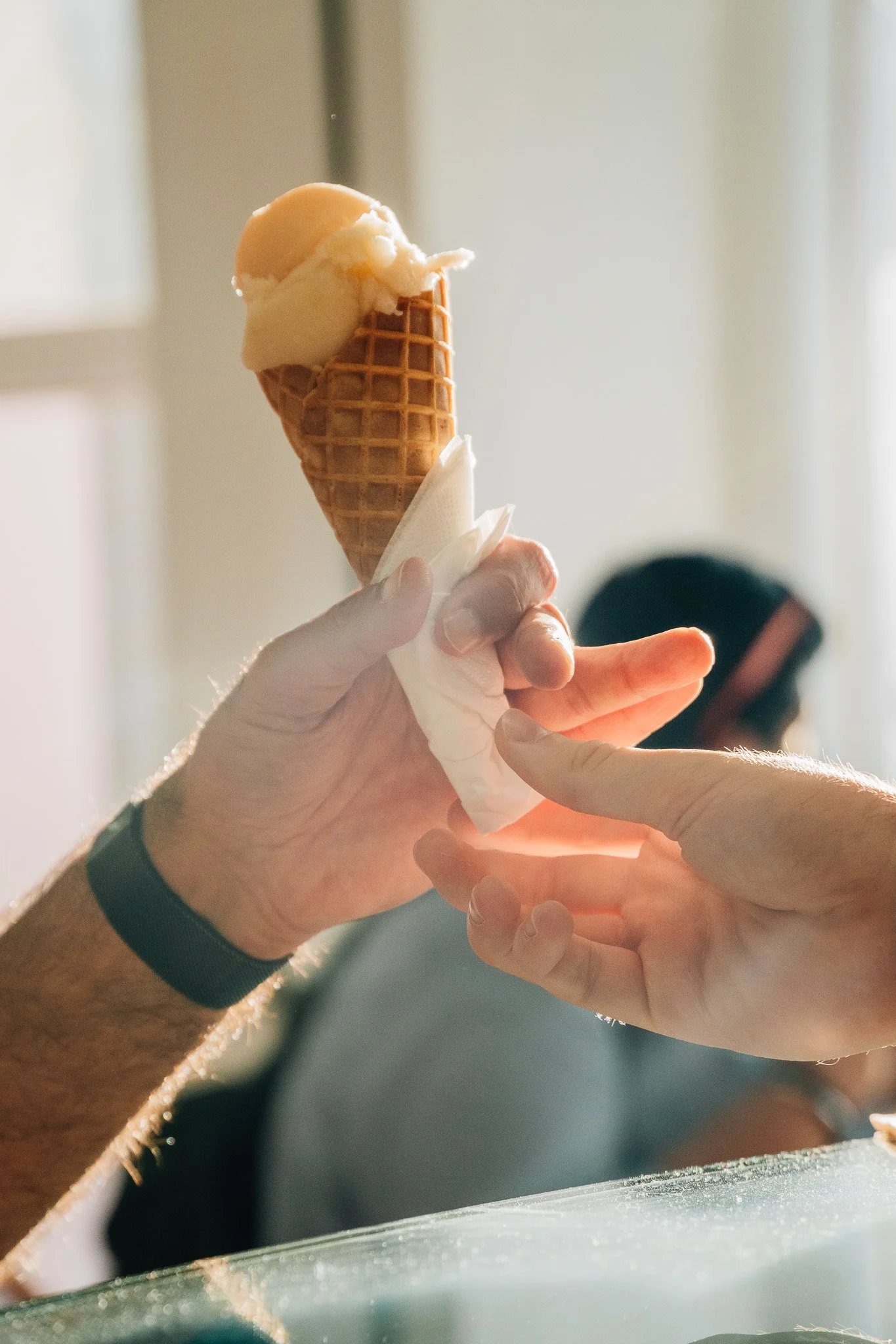 Close-up of a person holding an ice cream cone with two scoops of ice cream, one caramel-colored and the other vanilla, over a blurred background.