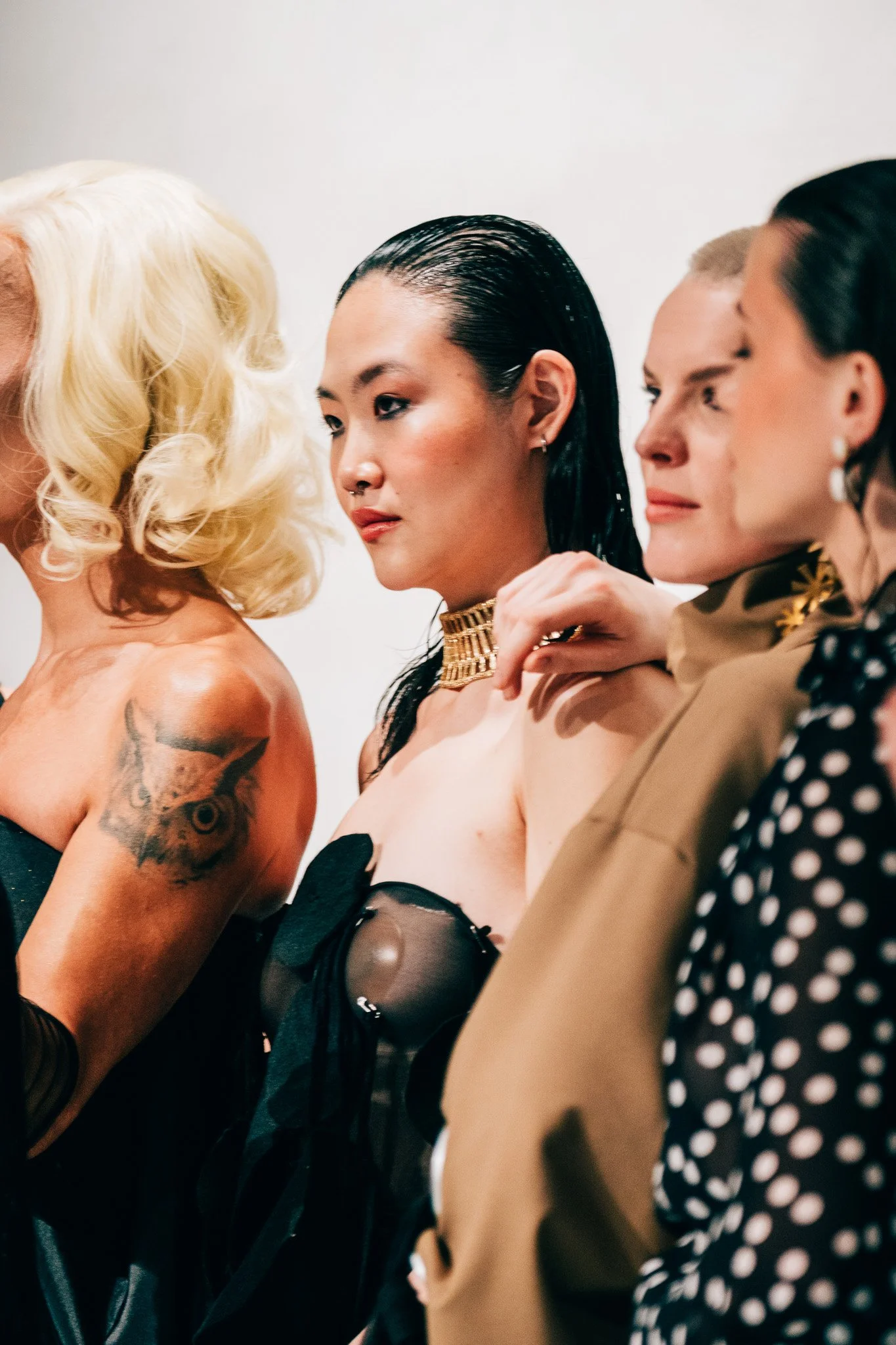 A close-up photo of four women standing in a row, their heads turned to the side, with various hairstyles and clothing styles visible.