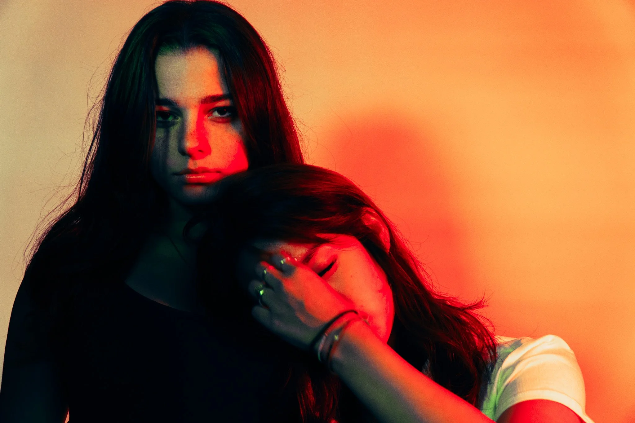 A woman with long dark hair looking at the camera, illuminated with red and green lighting, holding someone close in an embrace.
