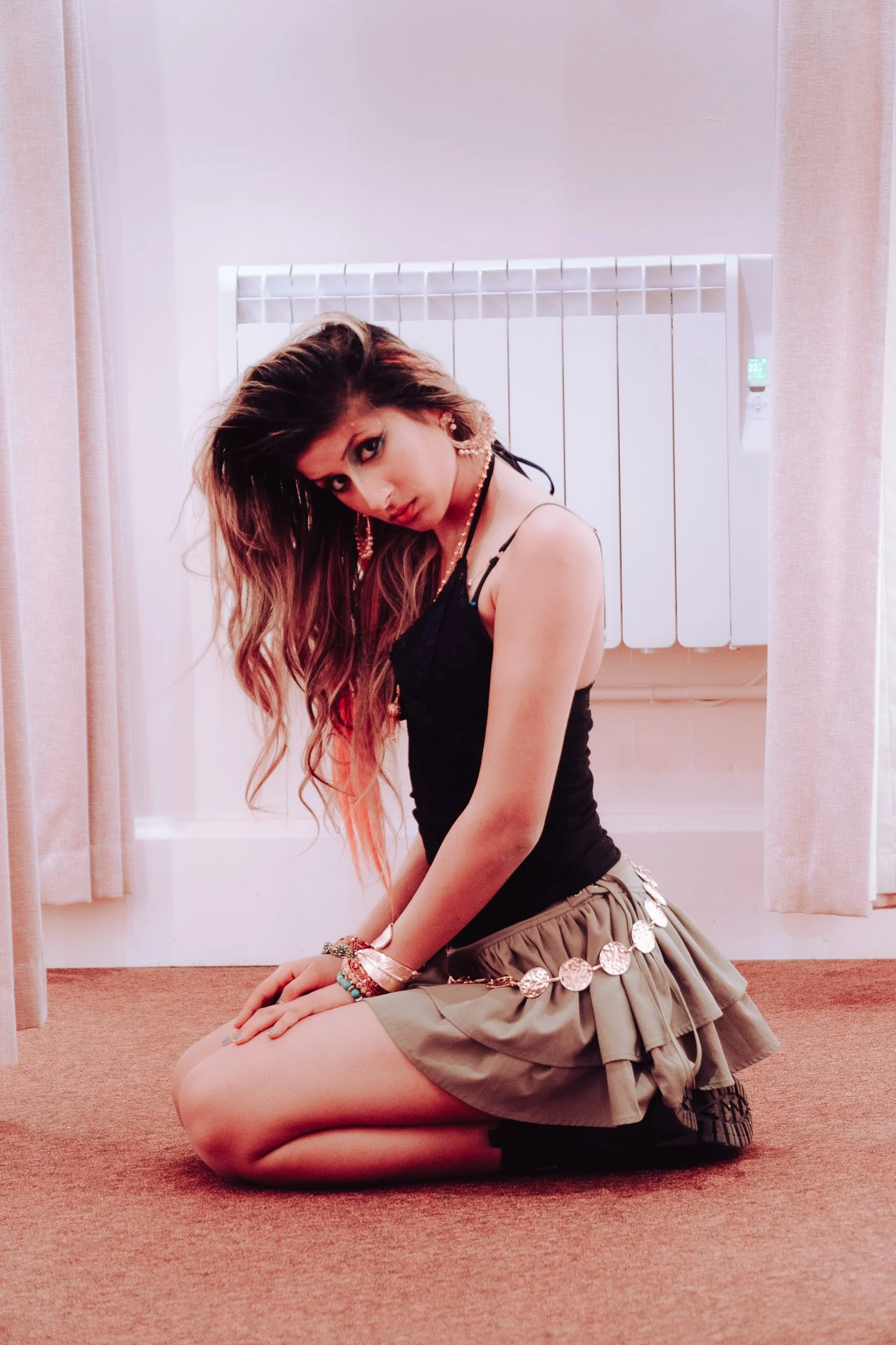 A young woman with long wavy hair dressed in a black tank top and beige pleated skirt, kneeling on a red carpet with beige curtains and a radiator in the background.