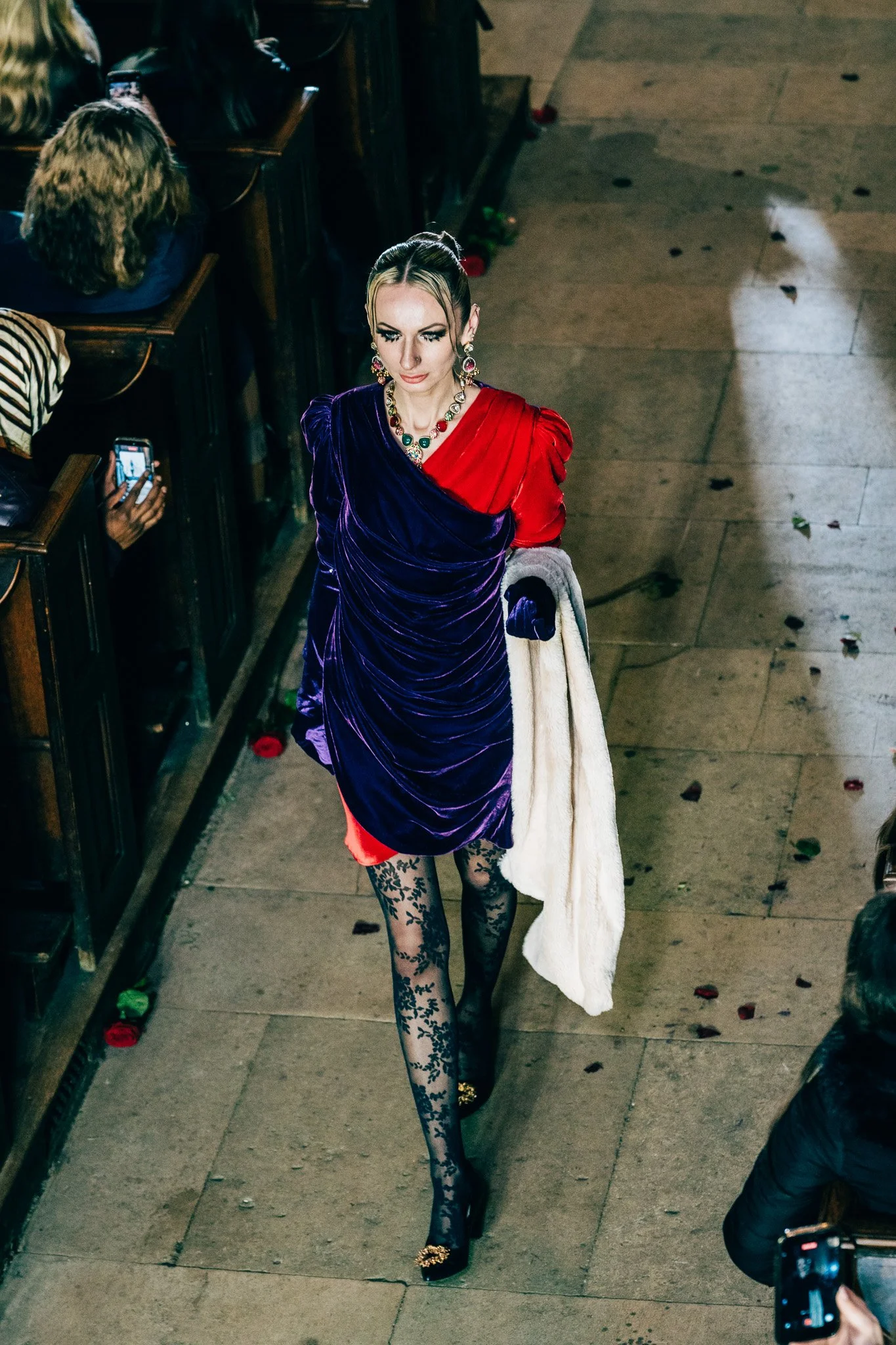 A woman in a colorful, elegant dress with tights and high heels walking on a sidewalk, surrounded by scattered flower petals, with people around taking photographs.