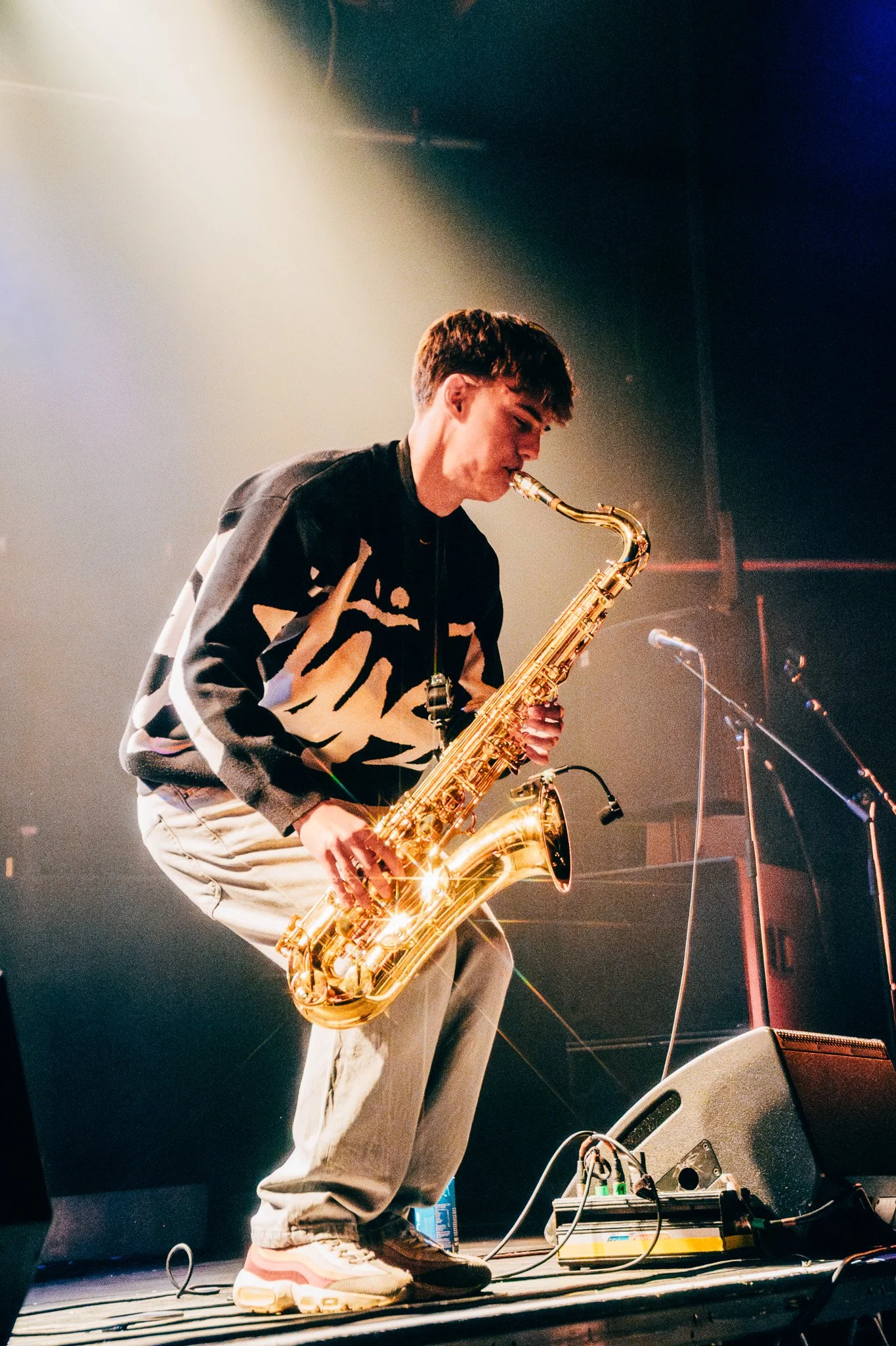 A young man playing a golden saxophone on stage, wearing a black and white patterned jacket, beige pants, and sneakers, with stage lights illuminating him.