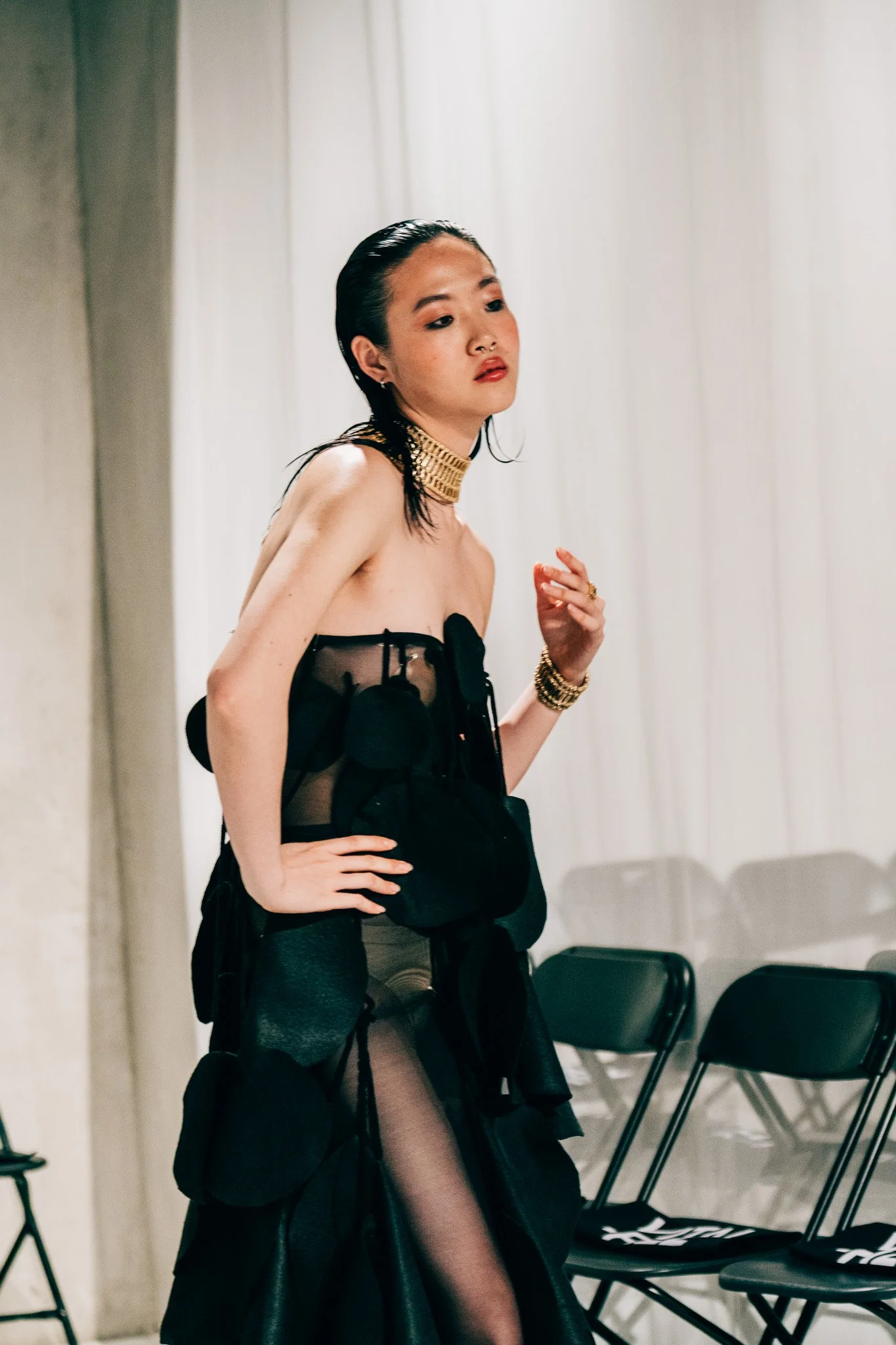 A woman with black hair and a nose ring wearing a black dress with sheer black stockings, accessories including a gold choker and bracelets, standing in front of a white curtain with black chairs behind her.