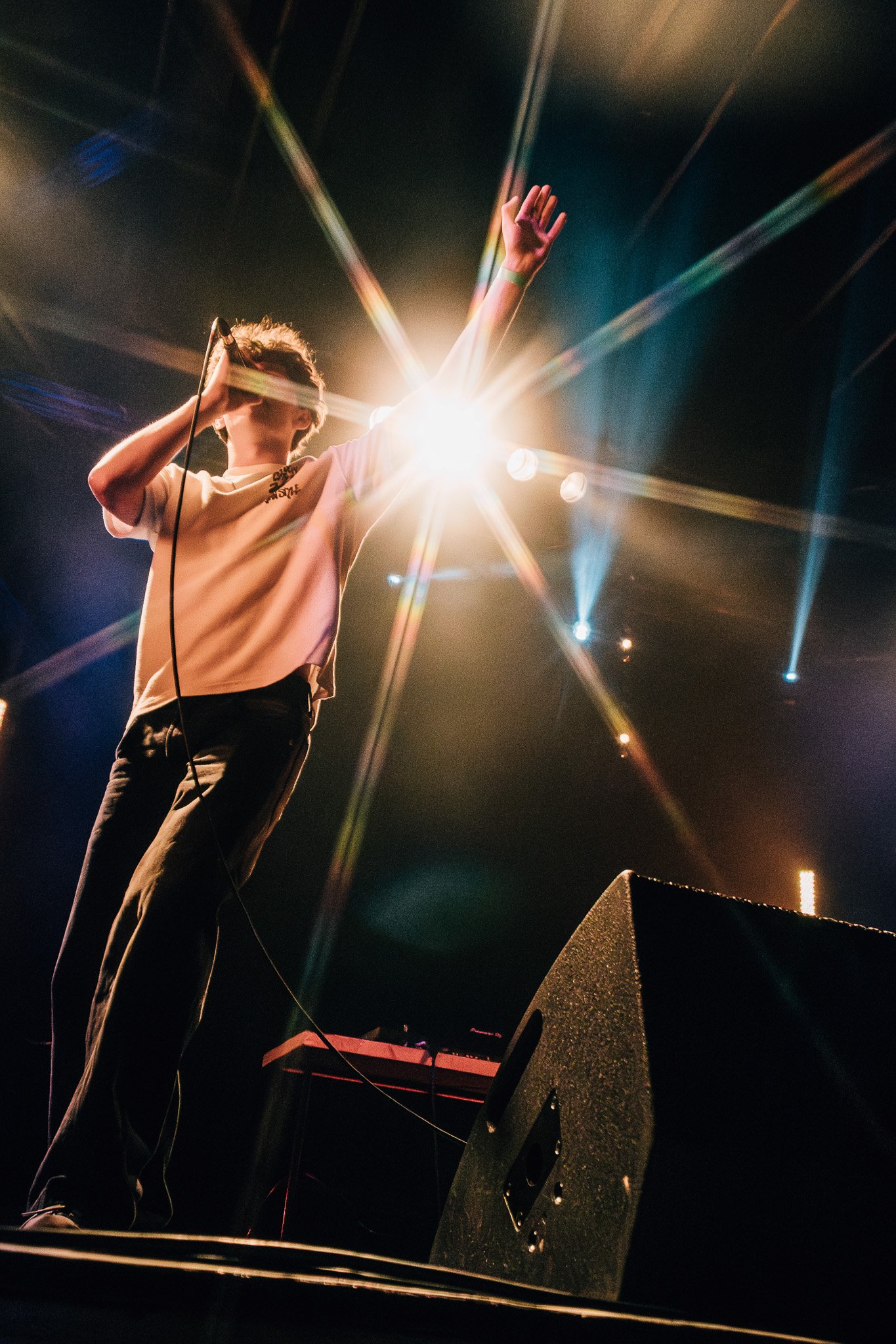 A person on stage performing with a microphone, with colorful stage lights and a spotlight behind them, wearing a beige T-shirt and dark pants.