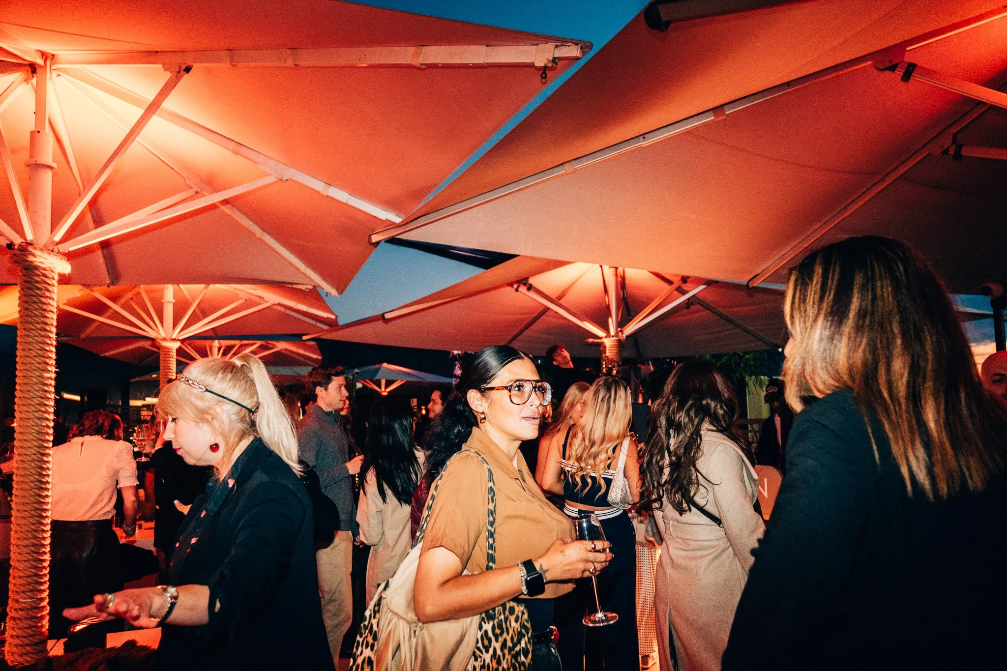 A crowd of people gathered outdoors at night under large orange umbrellas, some holding drinks, engaging in conversation, wearing fashionable clothing.