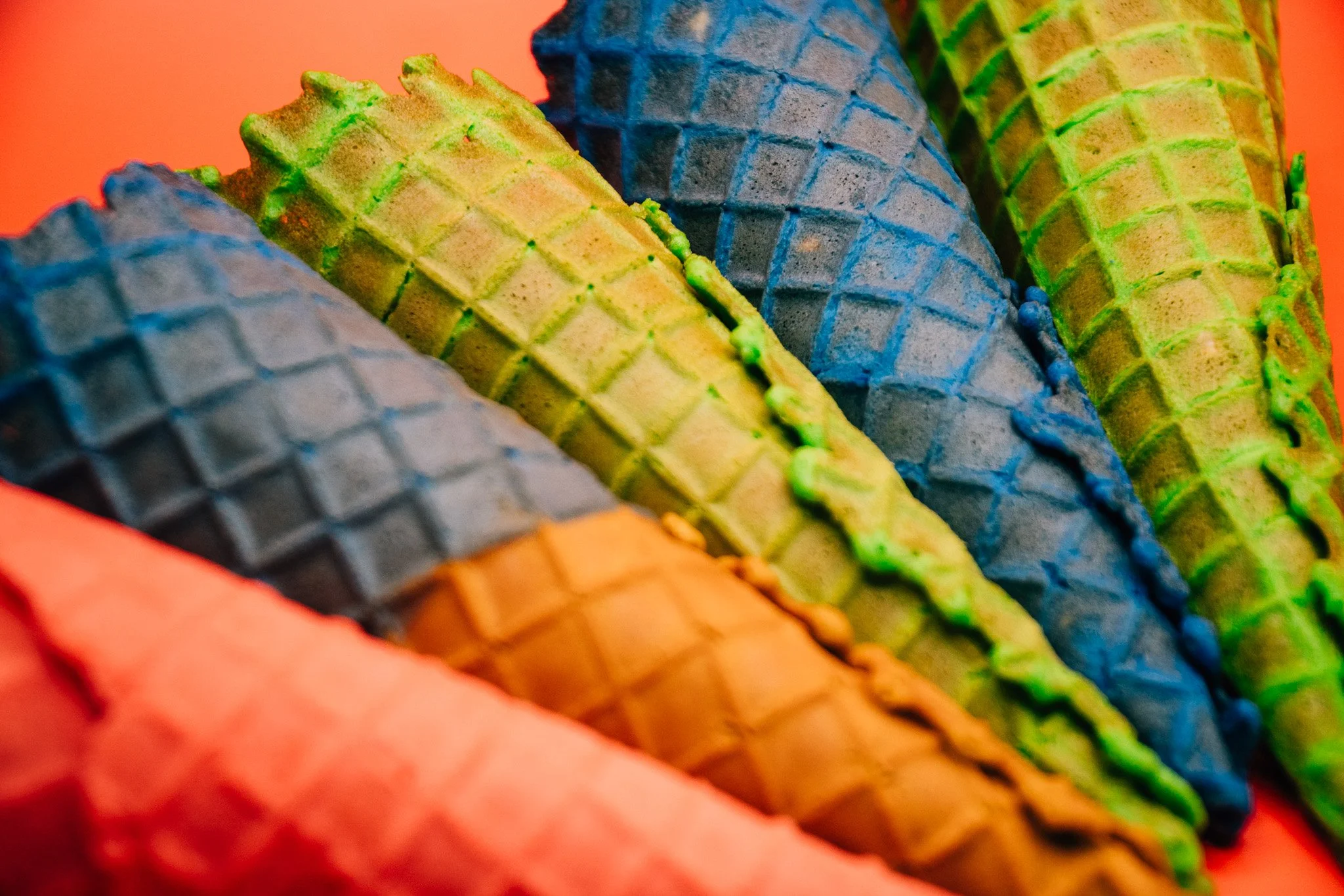 Close-up of vibrantly colored ice cream cones in bright orange container, with a mix of blue, green, yellow, and orange waffle cone patterns.