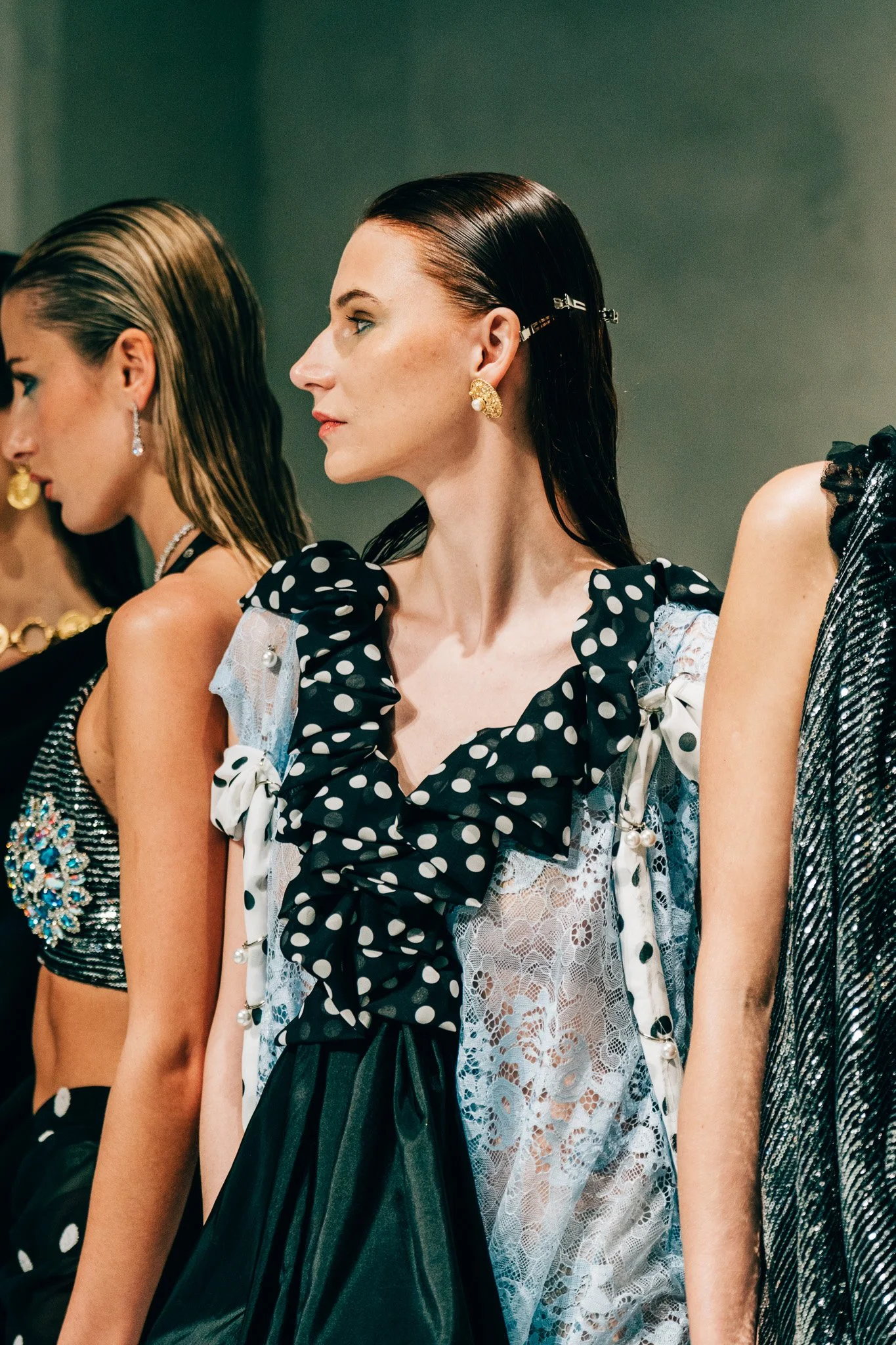 Three women with slicked-back hair and bold jewelry, wearing fashionable clothing, standing in a row.