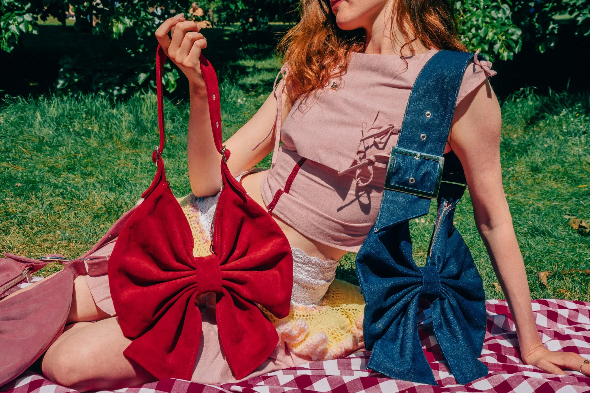 A woman with red hair sitting on a blanket outdoors, holding a red purse, wearing a pink top and checkered shorts, with trees in the background.