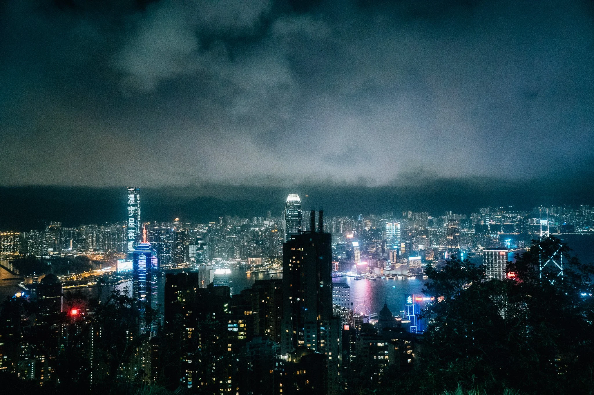 Nighttime city skyline with tall buildings illuminated, some with colorful lights, over a body of water with reflections. Dark clouds are in the sky.
