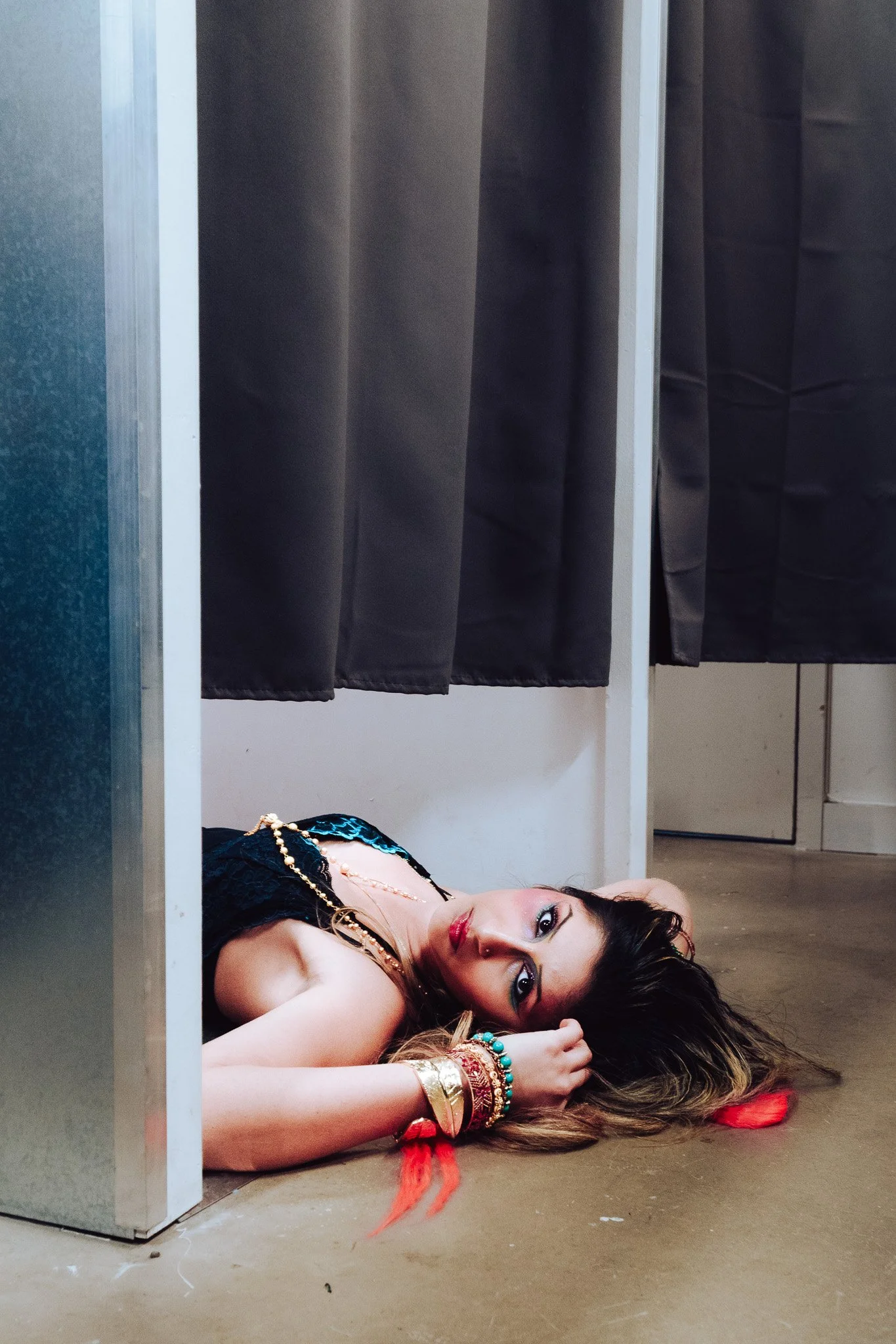 A woman lying on the floor with her head resting on her arm, wearing colorful jewelry and makeup, with a section of black curtains behind her.