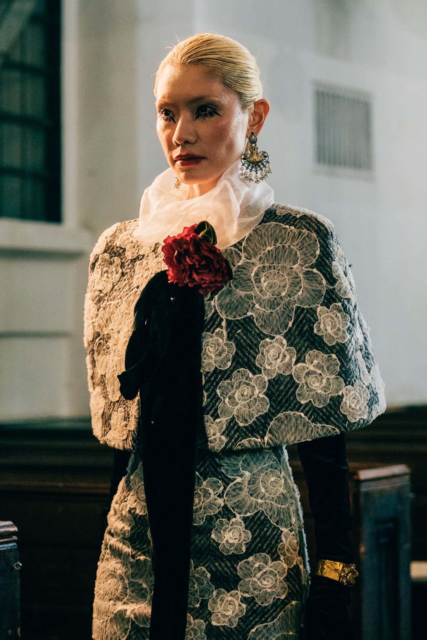 A woman with blonde hair, wearing ornate earrings, a white ruffled neck piece, a beige floral-patterned jacket, and black gloves with a gold bracelet, stands indoors with a serious expression.