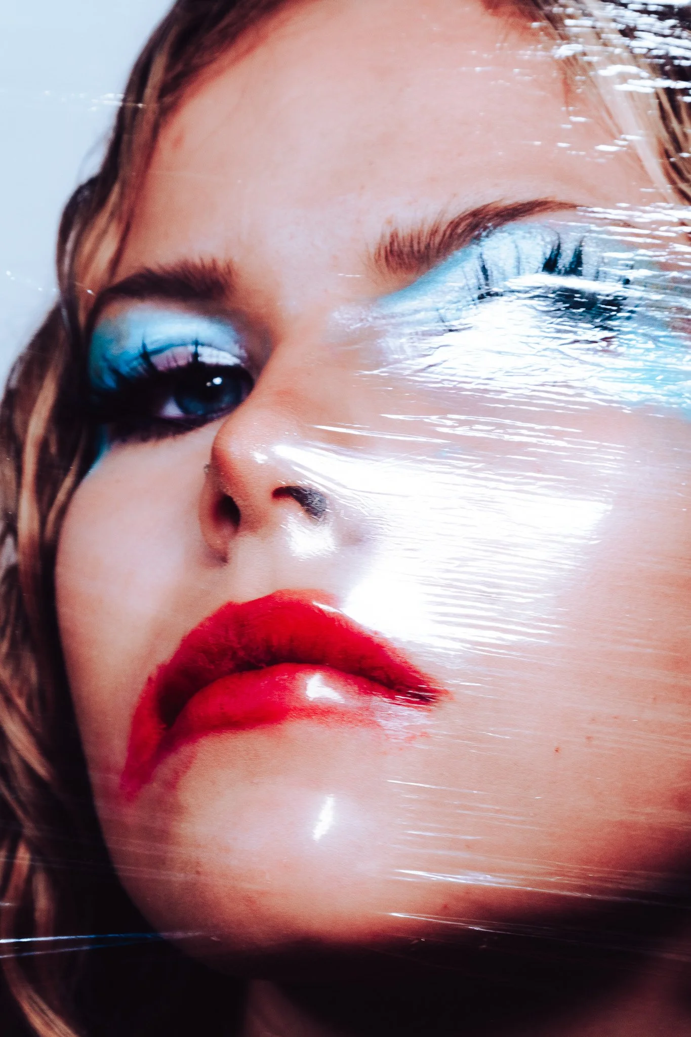 Close-up of a woman's face with bright red lipstick, blue eyeshadow, and long eyelashes. Her eyes are closed, and her face is partially covered with glossy plastic wrap.