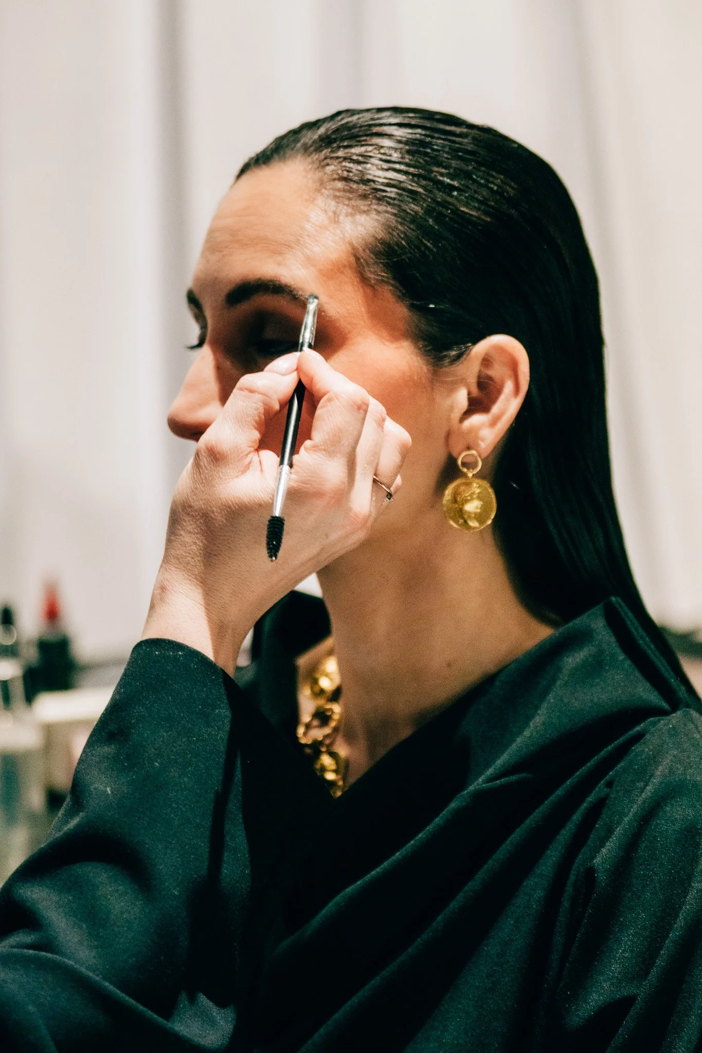 A woman with dark, slicked-back hair and gold earrings is getting her makeup done, specifically her eyebrows, by an artist using a makeup brush.