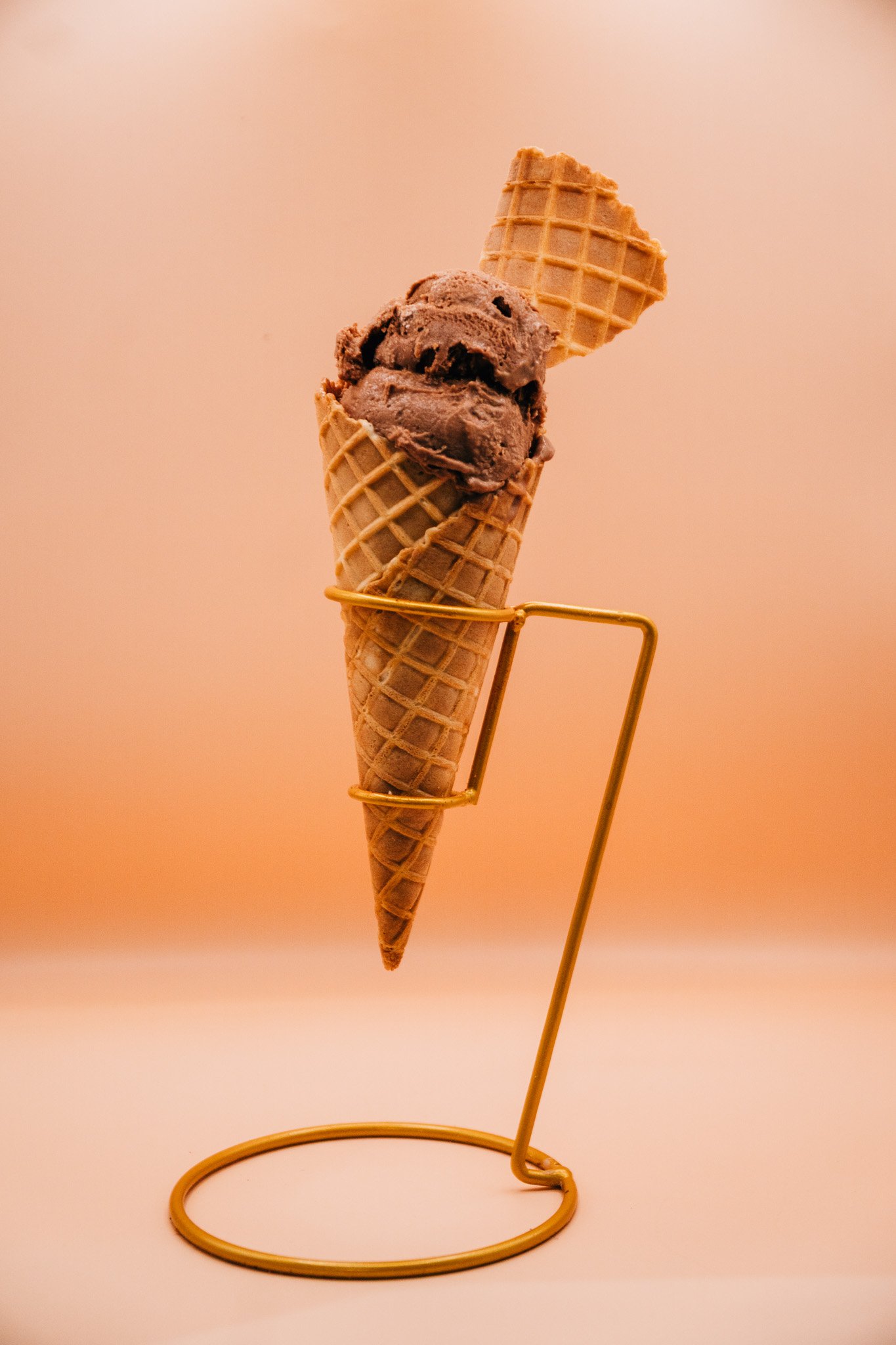Ice cream cone with two scoops of chocolate ice cream and a waffle cone, displayed on a gold wire stand against a soft peach background.