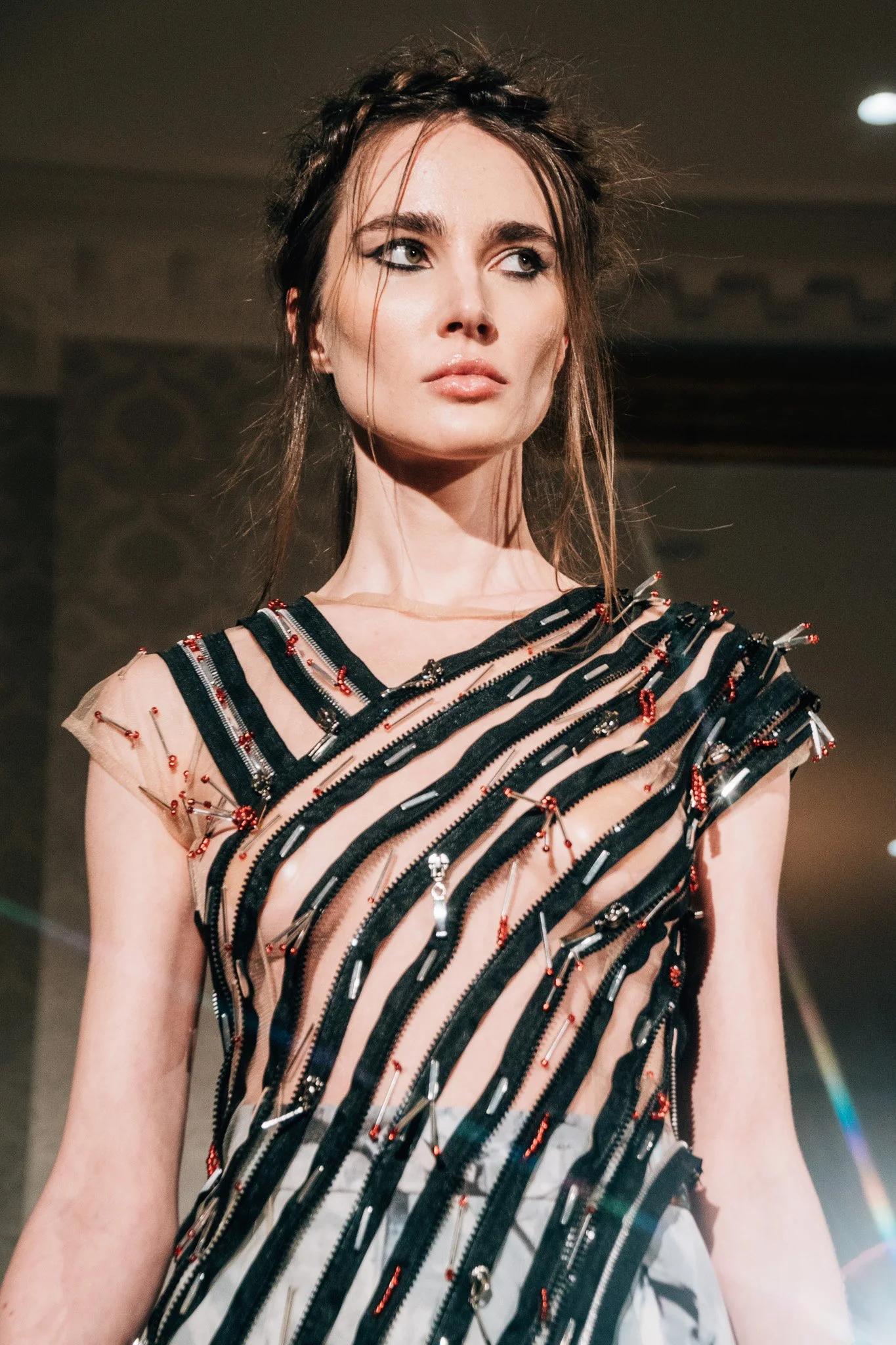 A woman with brown hair styled in a braided crown, wearing a dress made of clear material with black zippers and red embellishments, standing in an indoor setting with dim lighting.