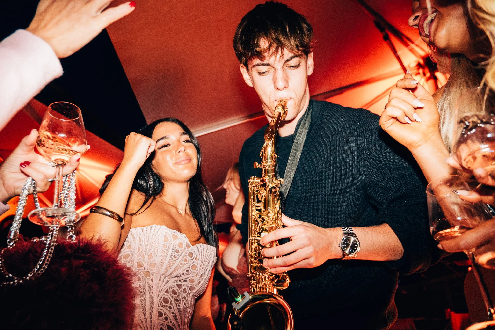 A young man playing a saxophone at a lively party with women dancing and holding drinks, in a dimly lit room with red lighting.