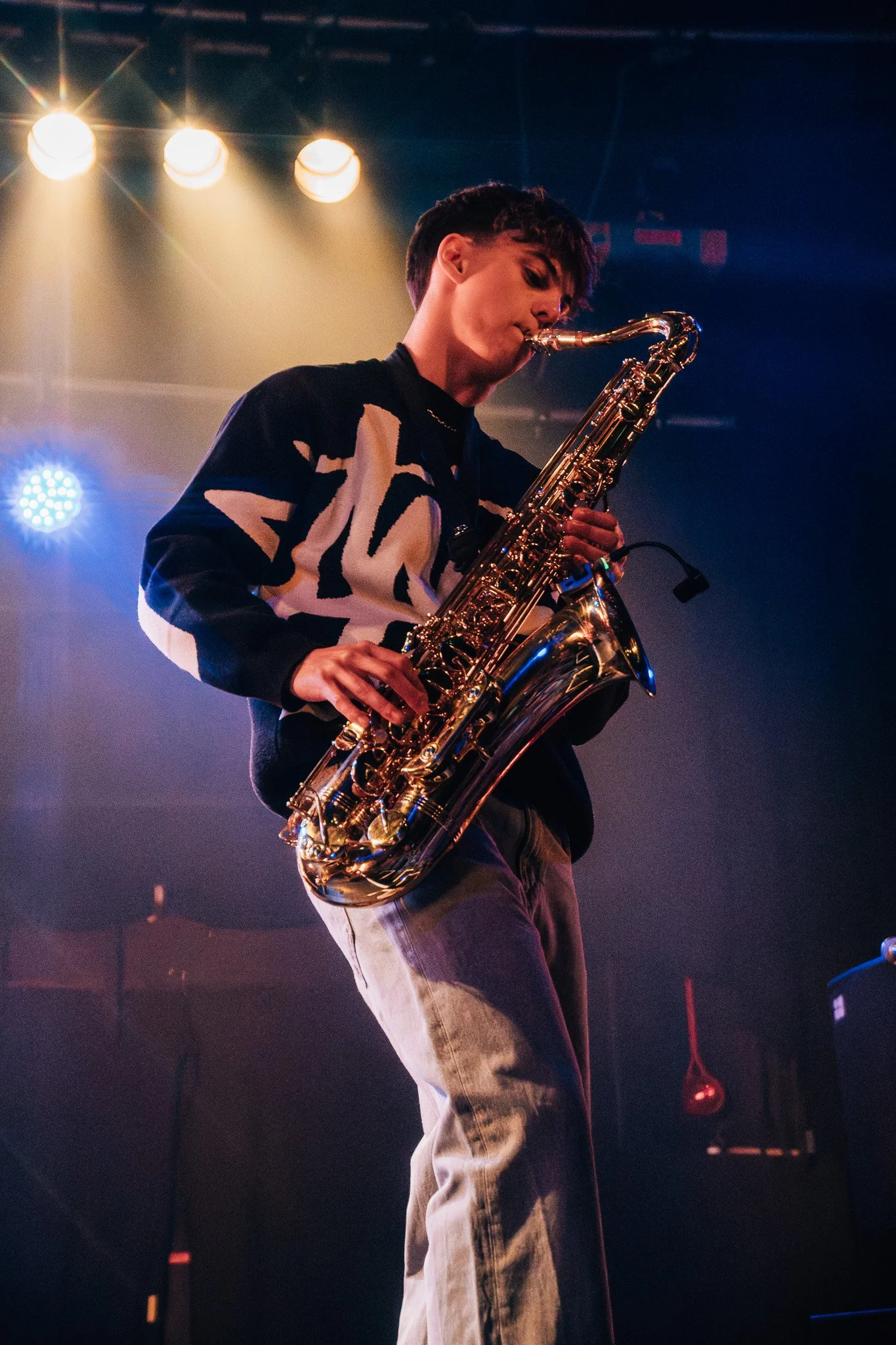 A young man playing a saxophone on stage with stage lighting and backdrops.