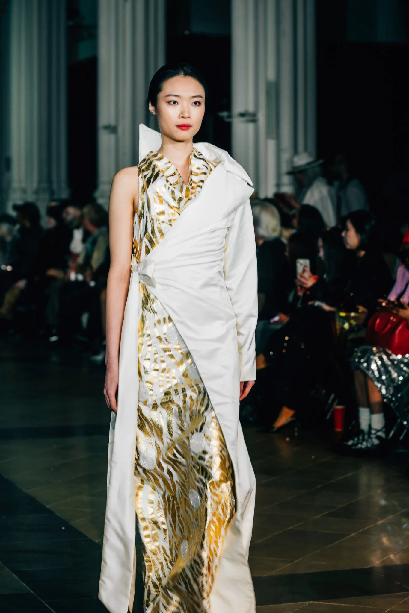 A female model walks on a runway wearing a white dress with gold and animal print details, surrounded by an audience at a fashion show.