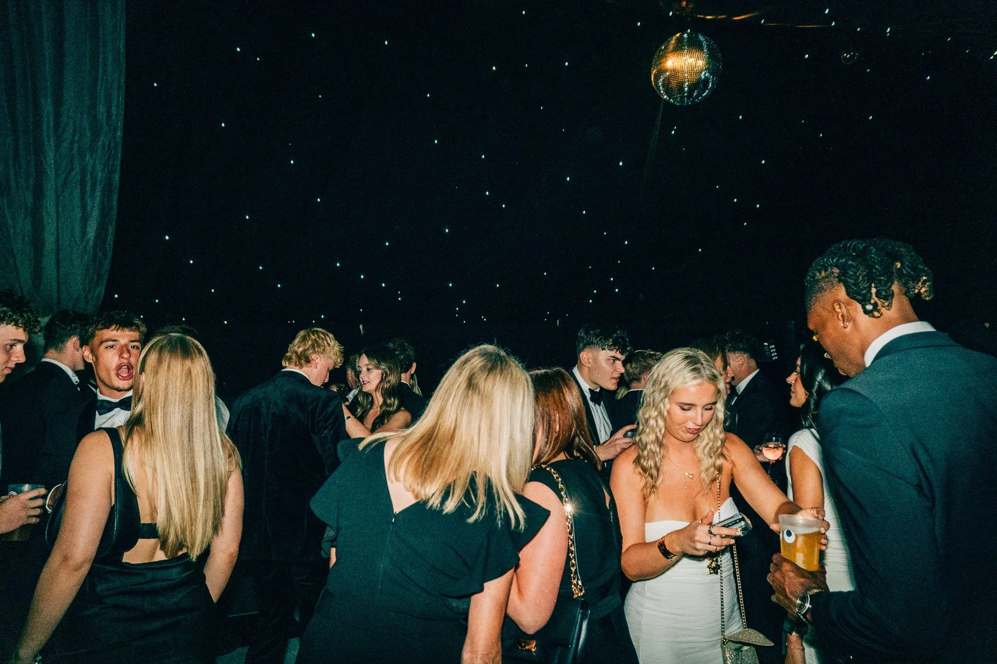 A group of young adults dressed in formal attire socializing and enjoying drinks at a party with a dark background and a disco ball overhead.