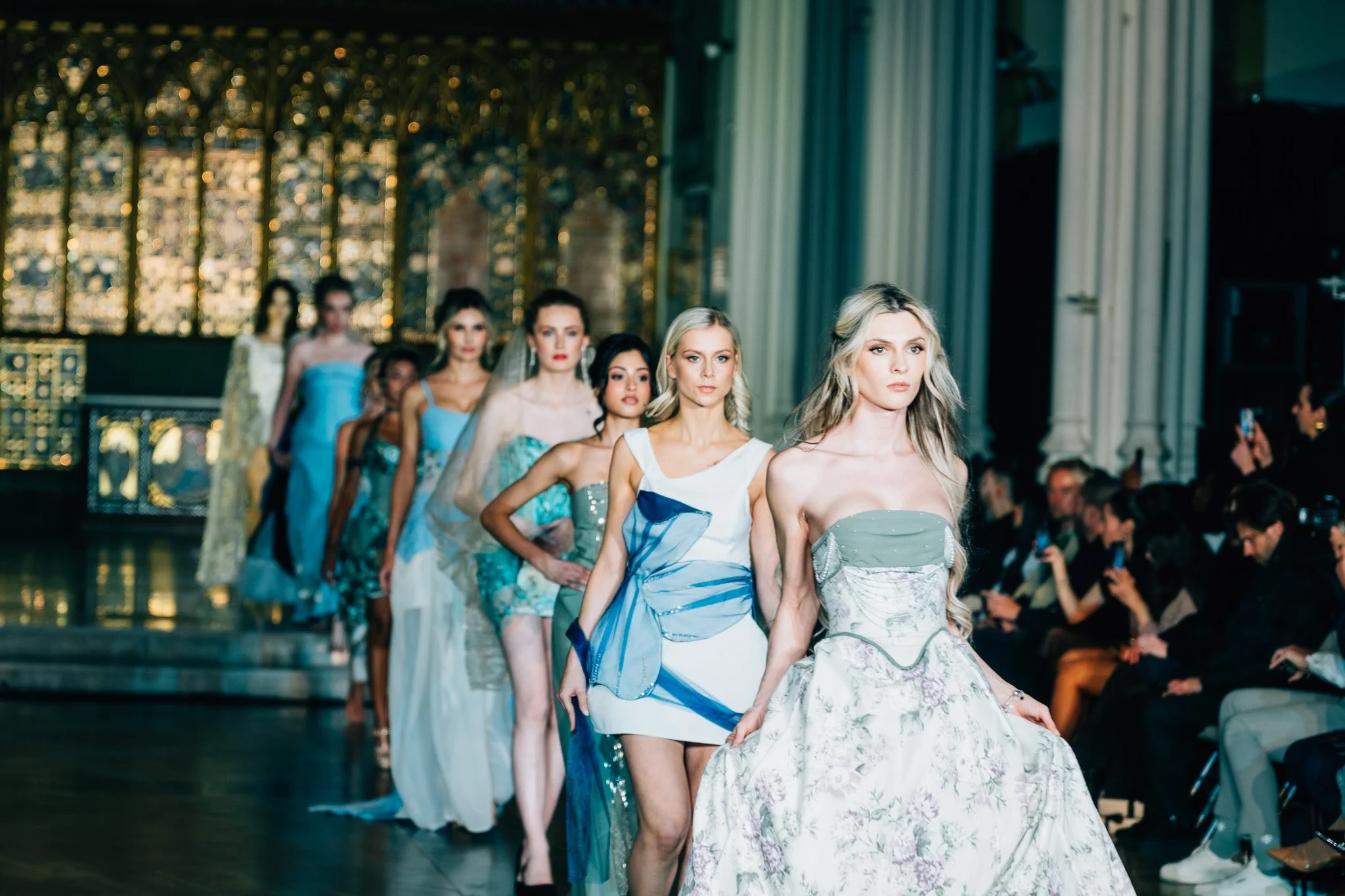 Models walk down runway during a fashion show in an ornate indoor venue with high ceilings, large windows, and decorative ironwork, wearing elegant dresses in pastel colors and floral patterns.