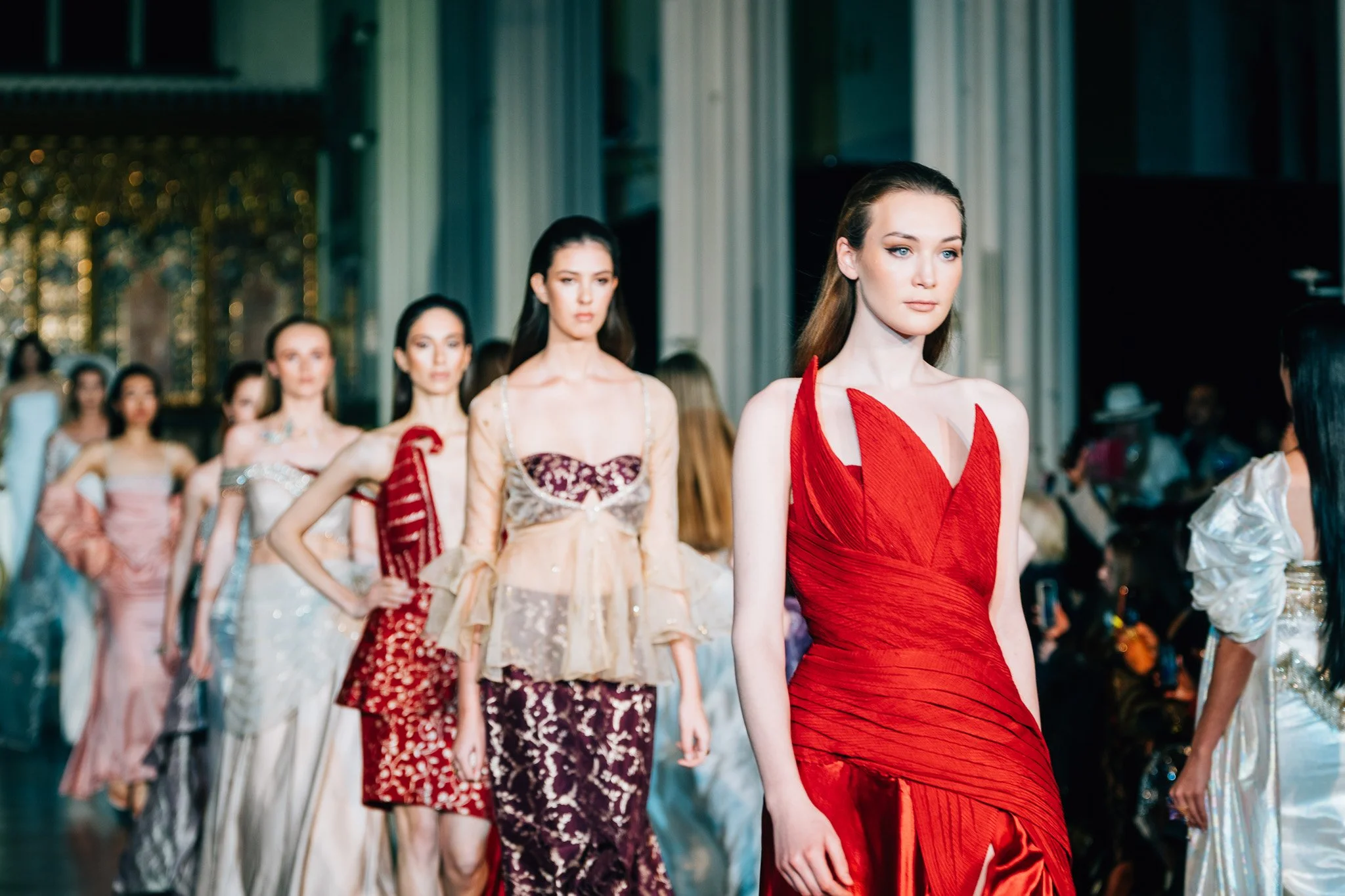 Models walk in a fashion show, with the lead model wearing a red dress and the others in various elegant outfits.