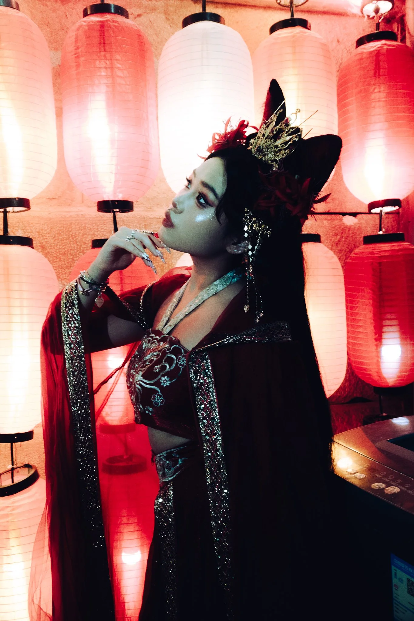 A woman dressed in traditional Asian attire with elaborate accessories, standing in front of illuminated red and white paper lanterns.