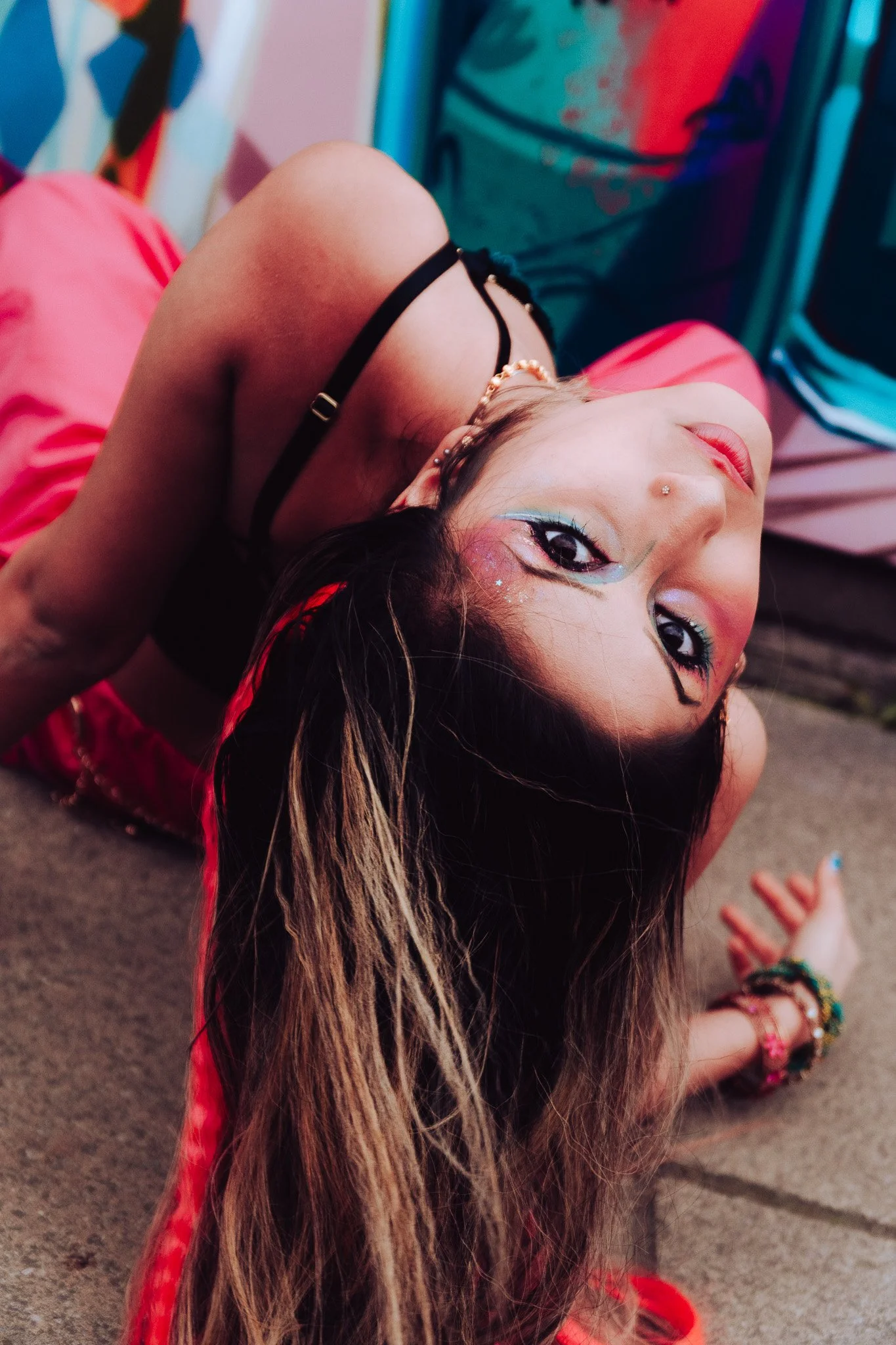 A woman with long, wavy hair lies on her back on a carpeted floor, with her head turned to the side. She has colorful makeup around her eyes, a nose piercing, and wearing a black top with thin straps. She is in a room with colorful, abstract wall art