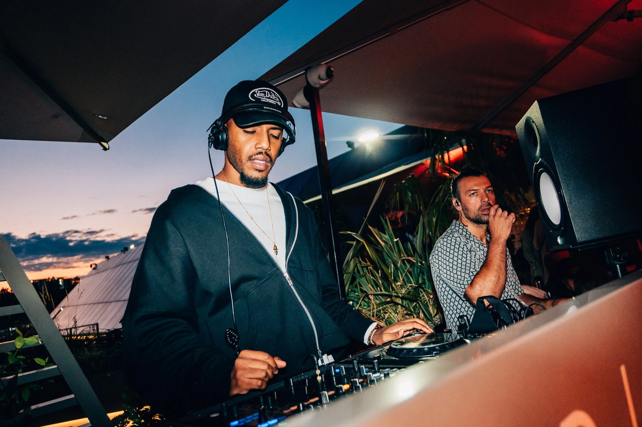 Two men are DJing at an outdoor event during sunset, one wearing a black Von Dutch cap and headphones, the other with hand near mouth, standing behind a DJ booth with equipment.