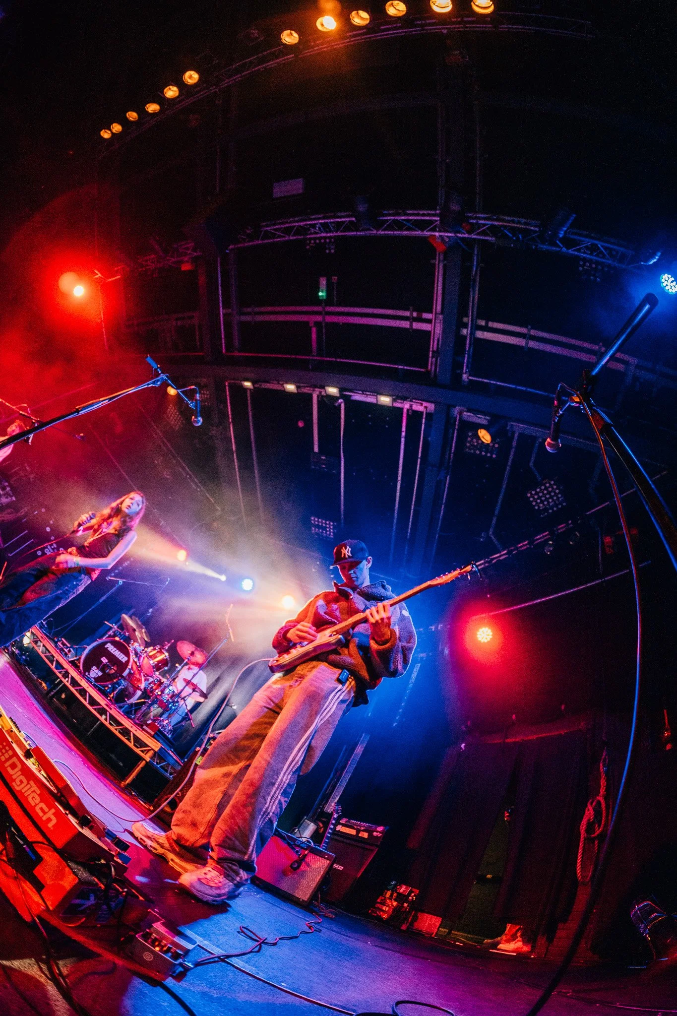 A band performing on stage with colorful lighting. A woman sings into a microphone, a man plays the guitar, and a drummer is visible in the background. The stage has various musical equipment and instruments.
