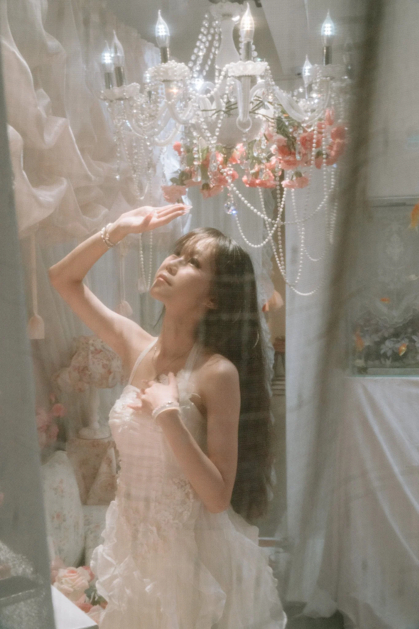 A woman dressed in a light pink and lace wedding gown is looking at herself in a mirror. She is inside a room with a decorative chandelier hanging from the ceiling, adorned with pink flowers and strings of pearls. The woman has long dark hair and is 