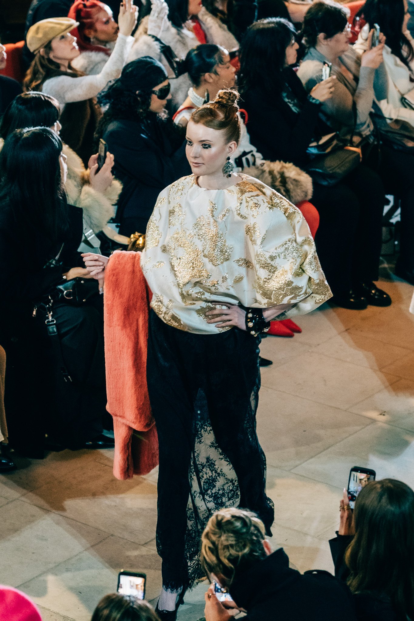 A woman standing confidently in front of an audience during a fashion presentation or event, dressed in a striking white and gold embroidered top and black lace bottoms, with her hair styled in an elegant updo.
