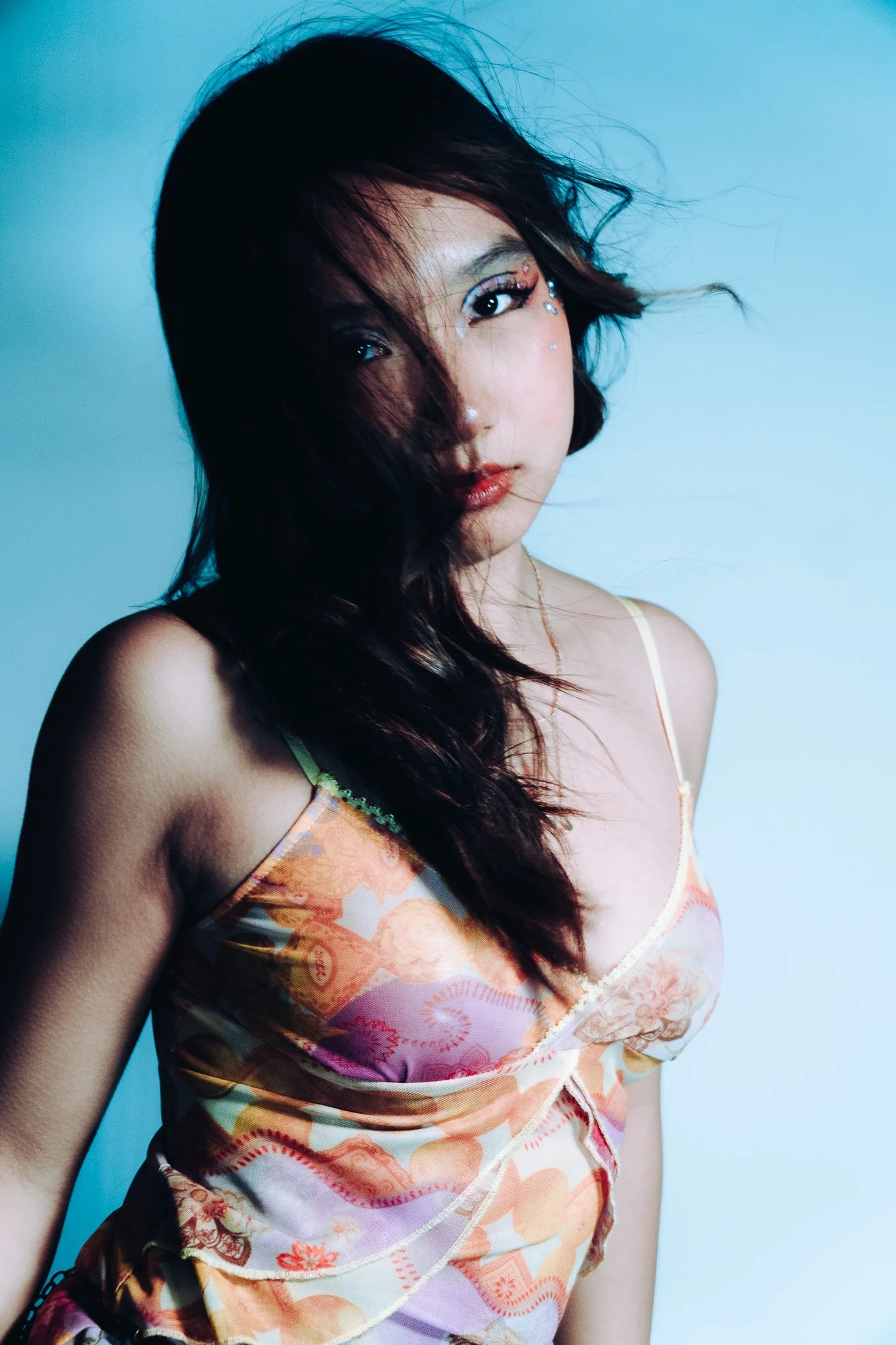 A woman with long dark hair and makeup, wearing a colorful patterned top, against a light blue background. Her hair partially covers her face, and she has decorative rhinestones near her eye.