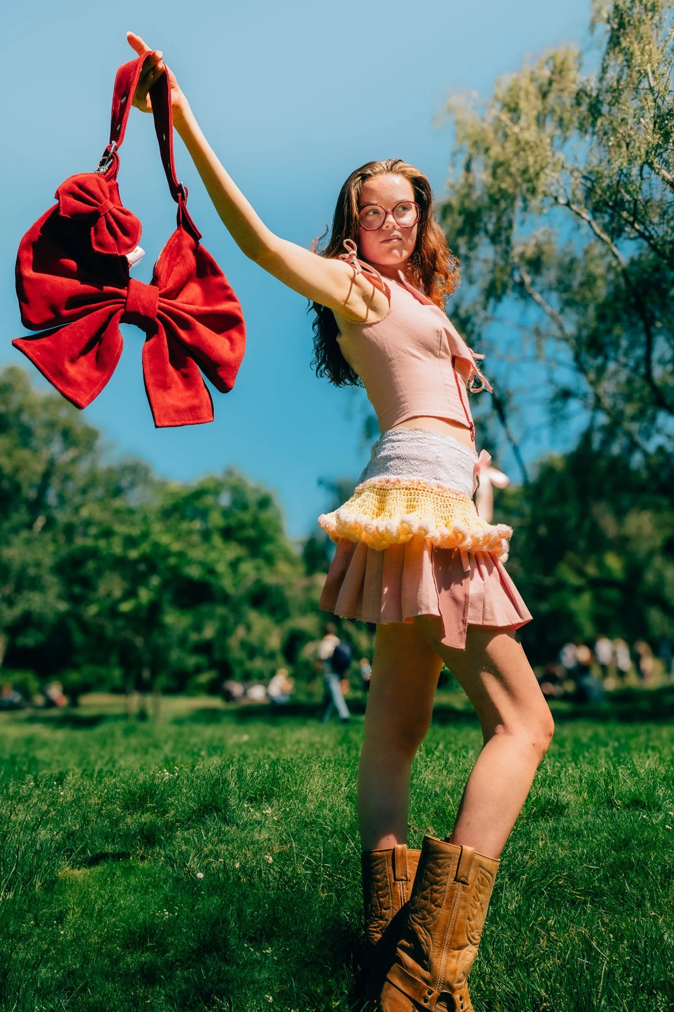 A young woman with red hair and glasses wearing a pink blouse with ruffled sleeves and a pink skirt with a yellow and orange ruffled hem, standing on grass in a park with trees and people in the background. She is holding a large red bow-shaped purse