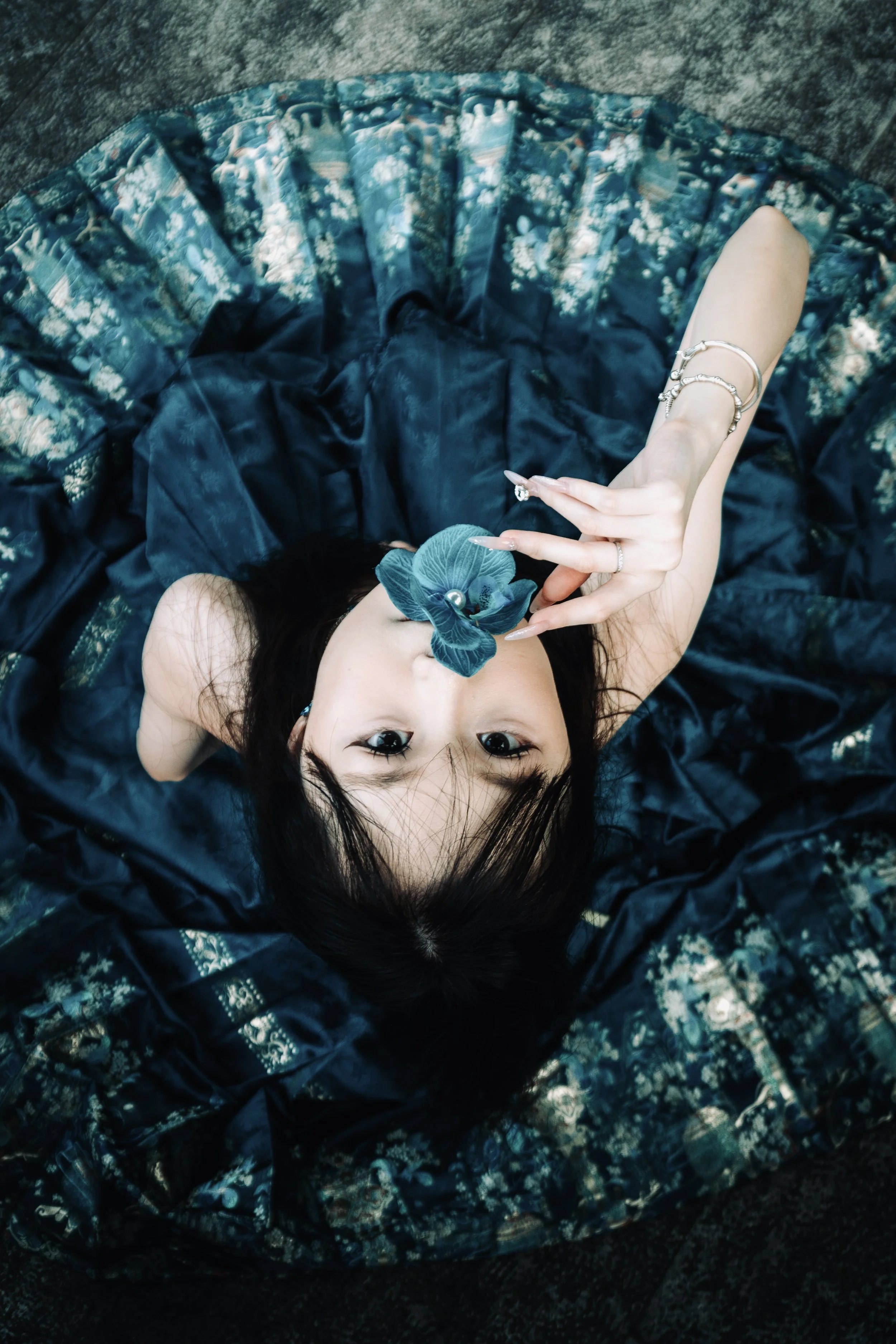 A woman lying on a floral-patterned blanket on the ground, holding a blue flower close to her face, wearing a dark-colored dress or skirt, with her arms adorned with silver jewelry.