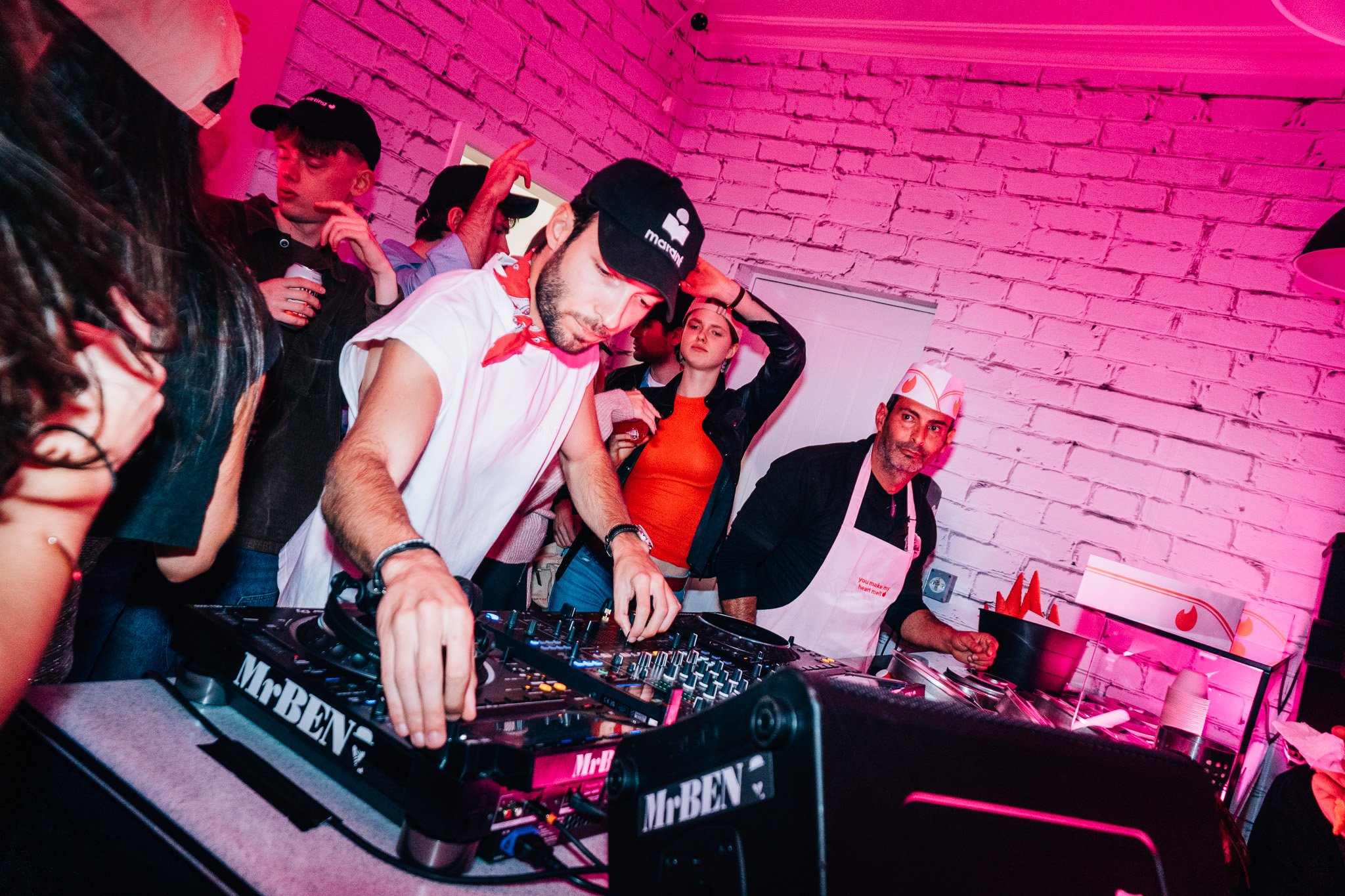 A DJ is performing at a party with pink lighting, surrounded by people, in a room with white brick walls. The DJ is wearing a cap and a white shirt, focused on the mixer, while others watch or dance.