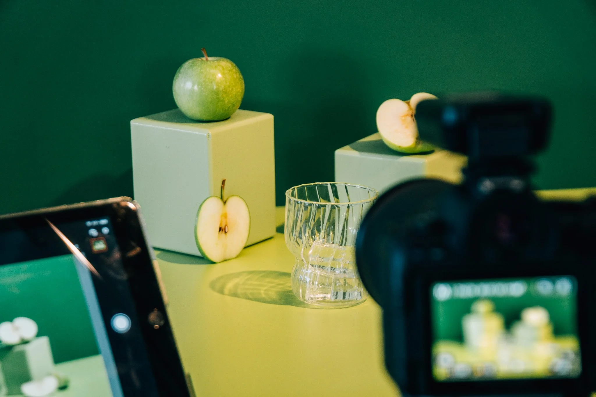 Green apple on a white pedestal, with half a sliced apple and a glass tumbler nearby, against a green background, with photo equipment partially visible.
