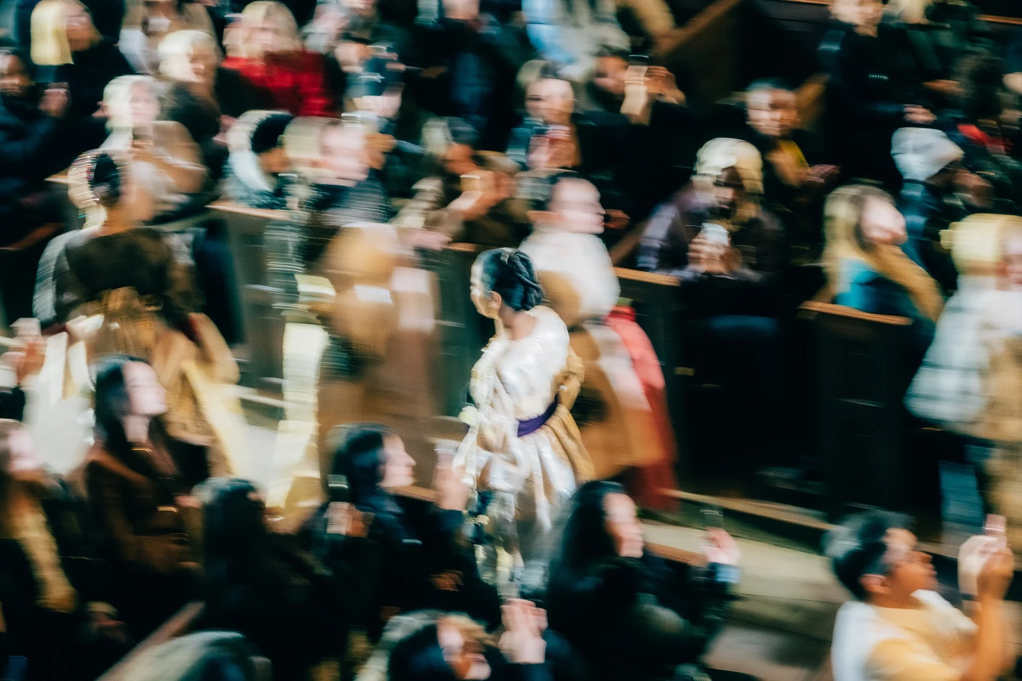 Blurry photo of a crowded indoor event with people sitting and standing, some dressed in formal attire.