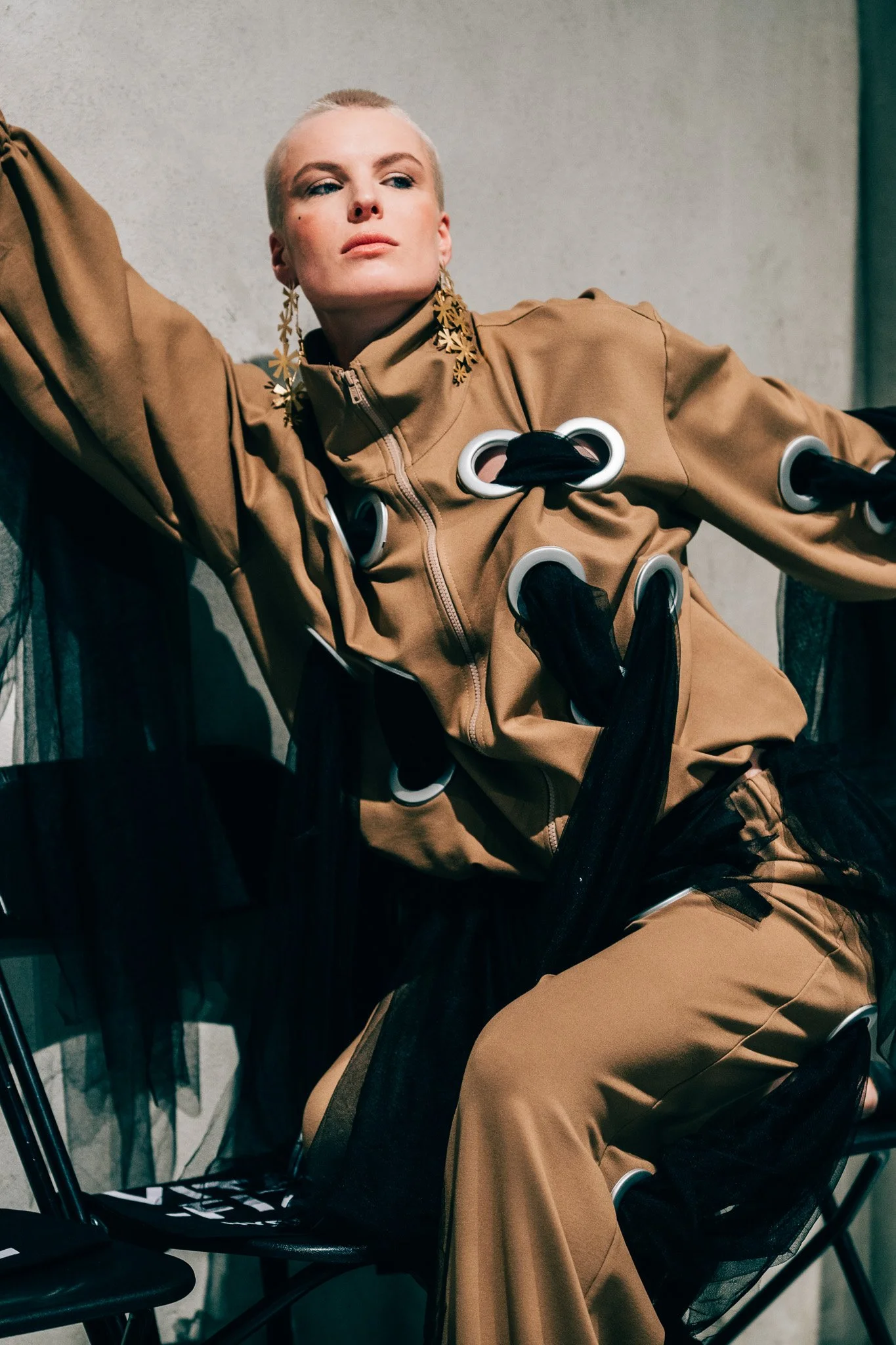 A woman with braided platinum blonde hair, wearing large star-shaped earrings and a tan jacket with circular metal accents, seated on a black chair against a plain wall.