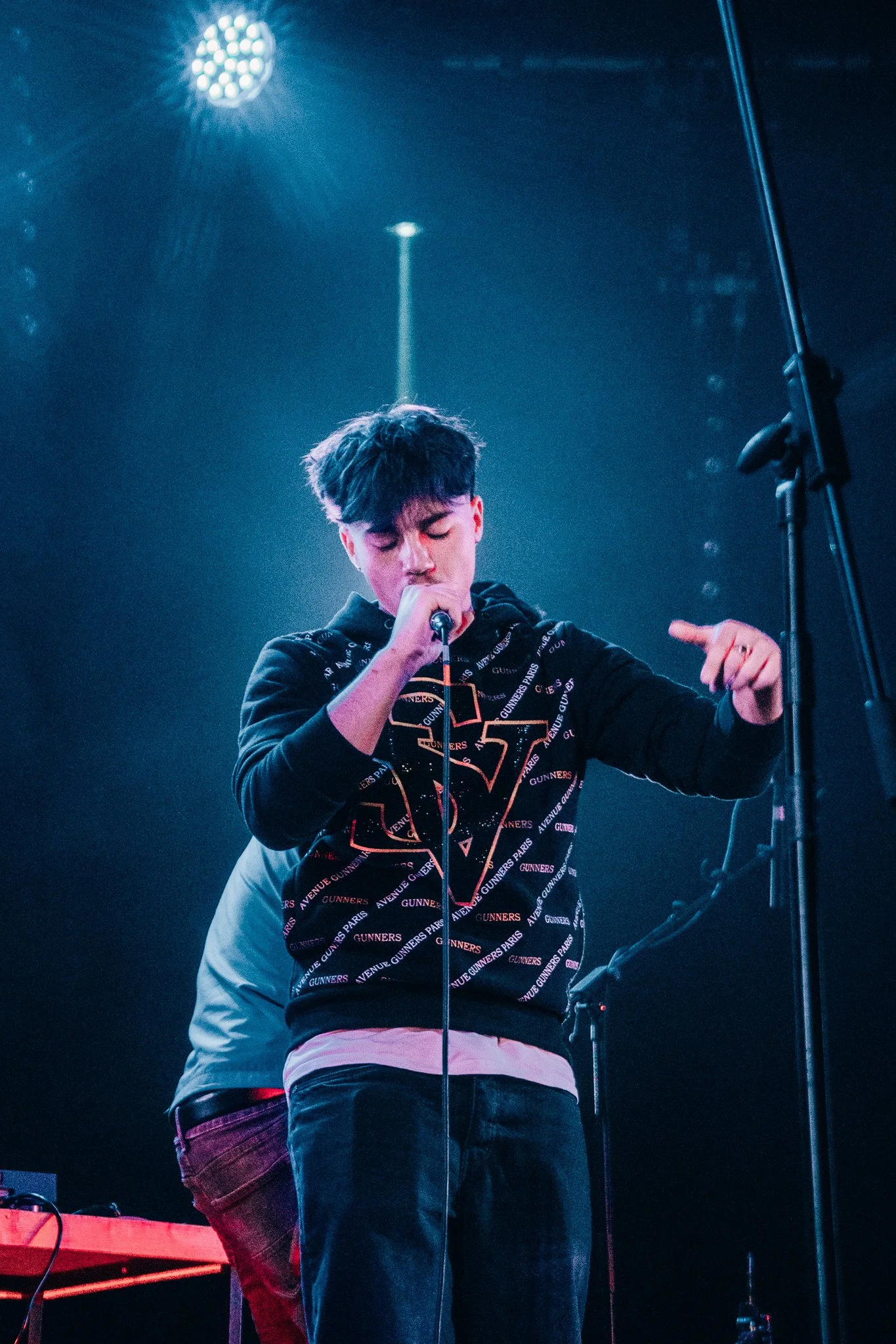 Young man singing into a microphone on stage, wearing a black hoodie with text and graphics, with stage lighting in the background.