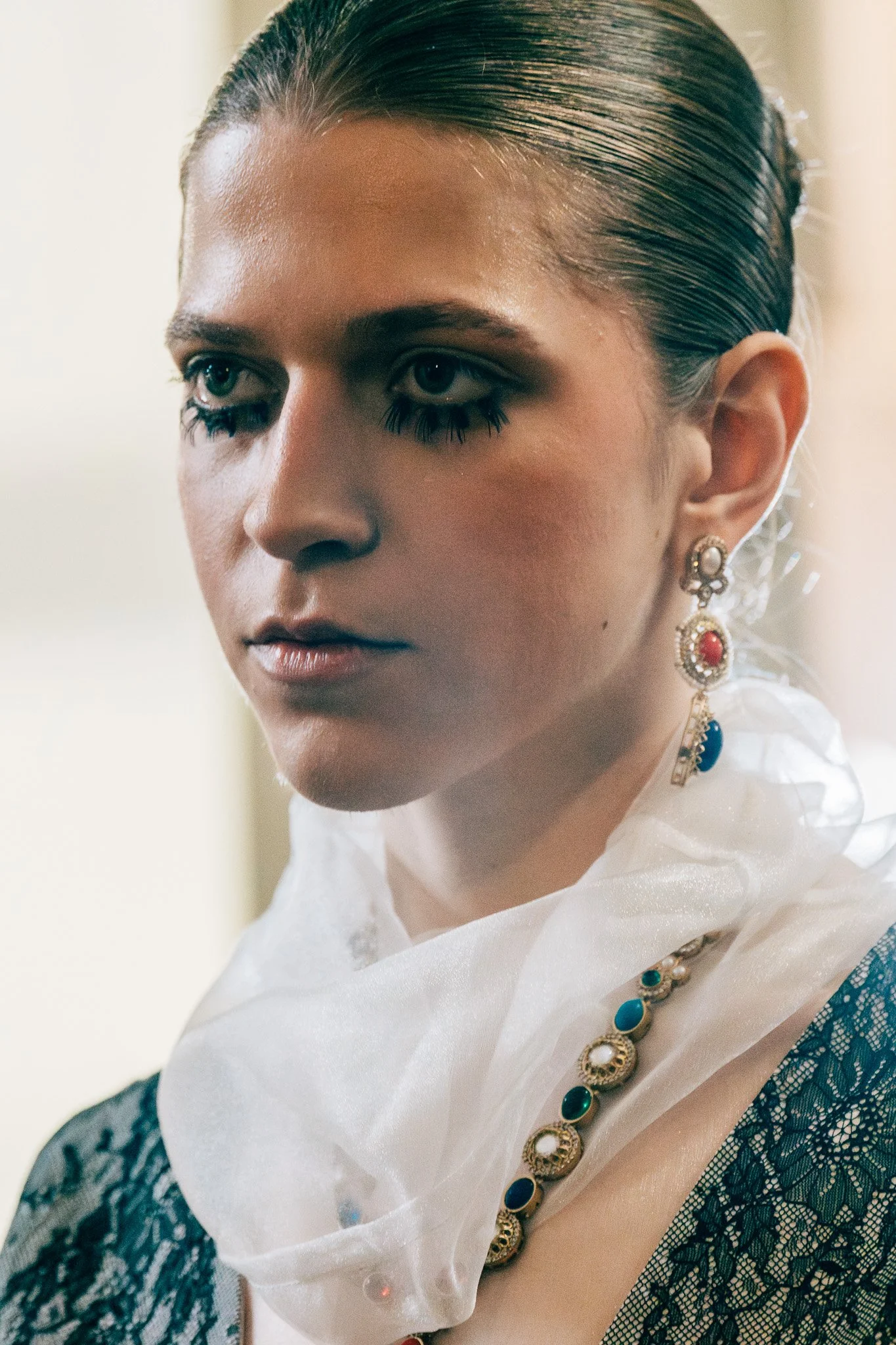 Close-up of a woman with wet hair, wearing large pearl and colored gemstone earrings and a black lace dress with a white satin shawl.