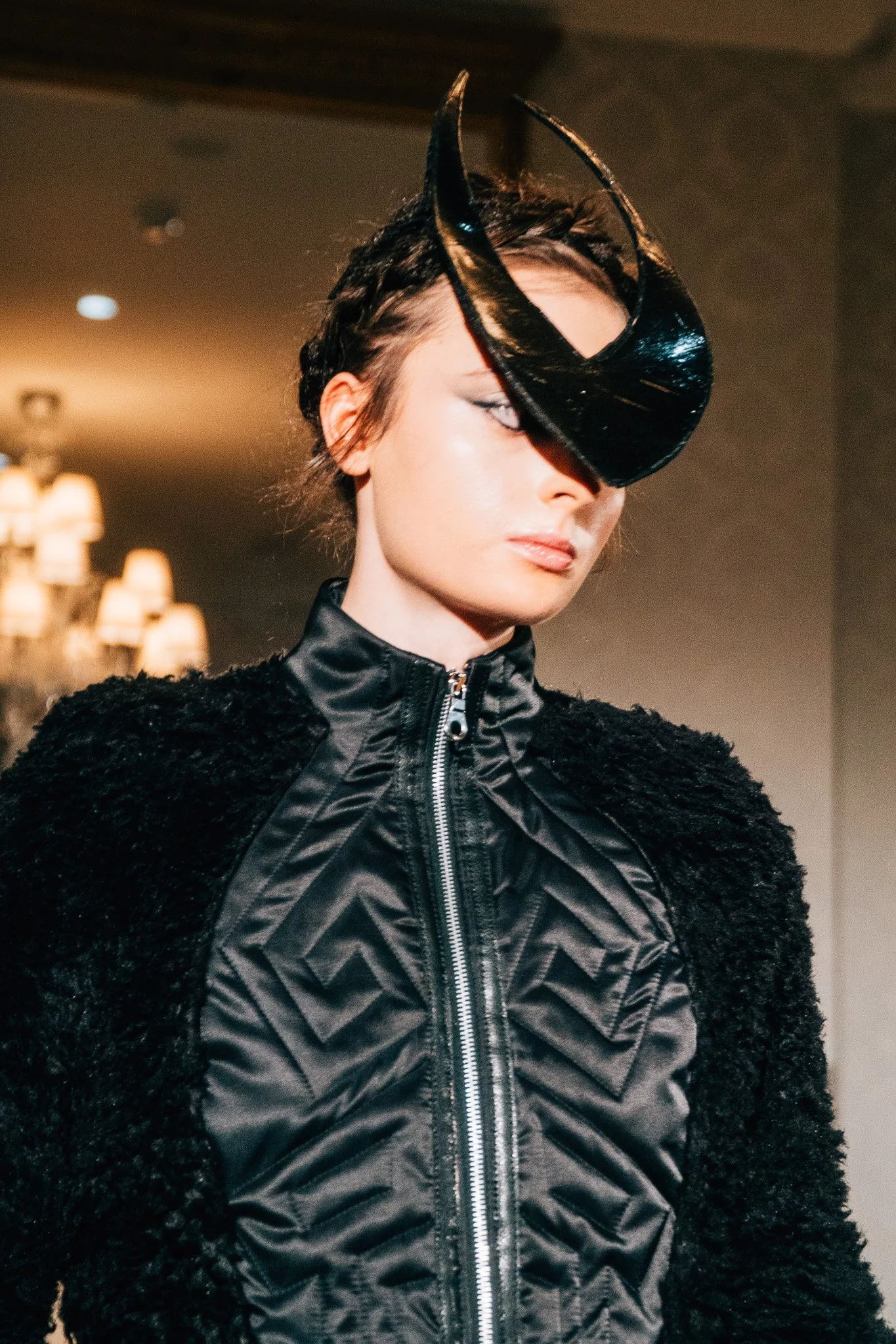 A model wearing a black hat with a large, curved brim, half covering her face, and a black zip-up jacket with a textured pattern and fluffy black sleeves, in an indoor setting with warm lighting.