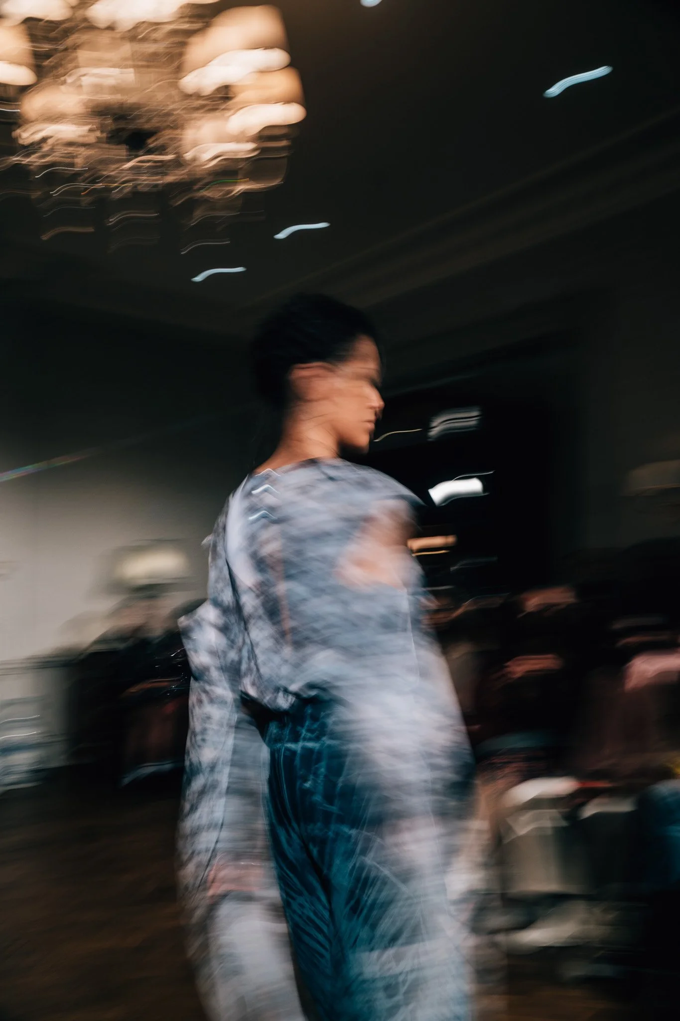 A woman with dark hair wearing a tie-dye shirt and jeans stands inside a dimly lit room with a large chandelier overhead, creating a blurred motion effect.